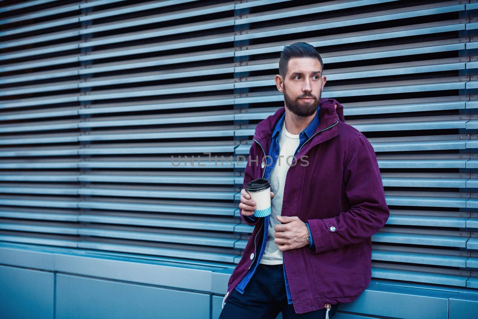 Outdoor fall or winter portrait of handsome hipster man with beard, white t-shirt, blue shirt and maroon jacket holding cup of hot coffee. Ribbed urban wall background.