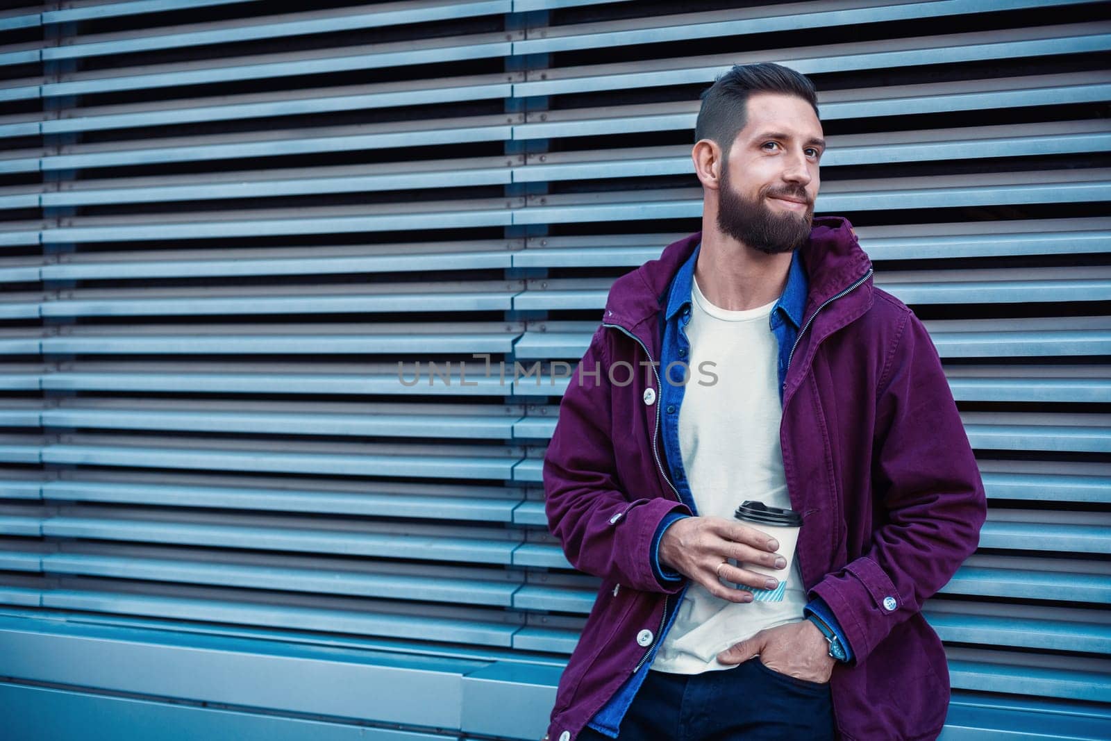 Outdoor fall or winter portrait of handsome hipster man with beard, white t-shirt, blue shirt and maroon jacket holding cup of hot coffee. Ribbed urban wall background.