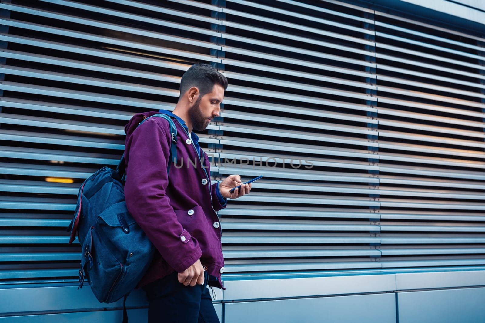 Confident bearded man holding in and mobile phone while standing near street wall in sunny summer evening. Young fashionable hipster guy using cell telephone while posing outdoors near copy space area