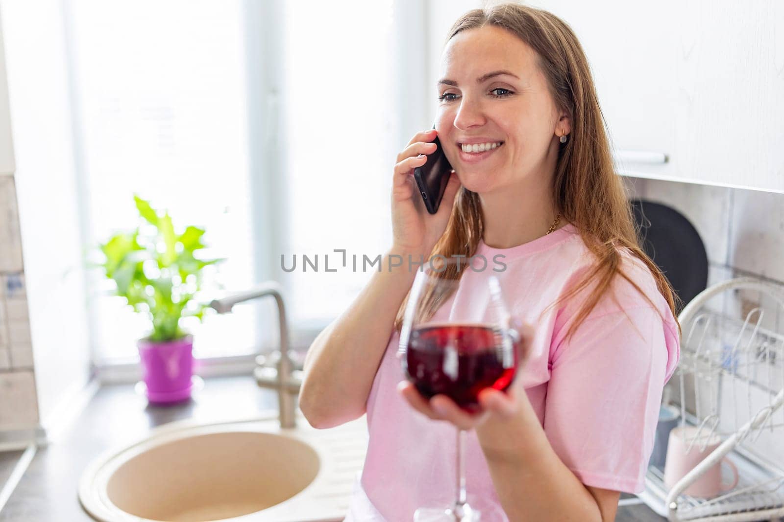 Happy relaxed young woman standing in kitchen with glass of red wine and using her smartphone.