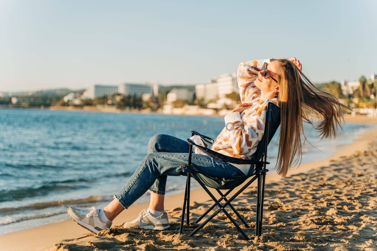 Beautiful young girl in cozy sweater and sunglasses stretching while sitting on foldable chair on winter beach sand. Woman enjoying autumn ocean fall sunset view while wind blowing her hair by Ostanina