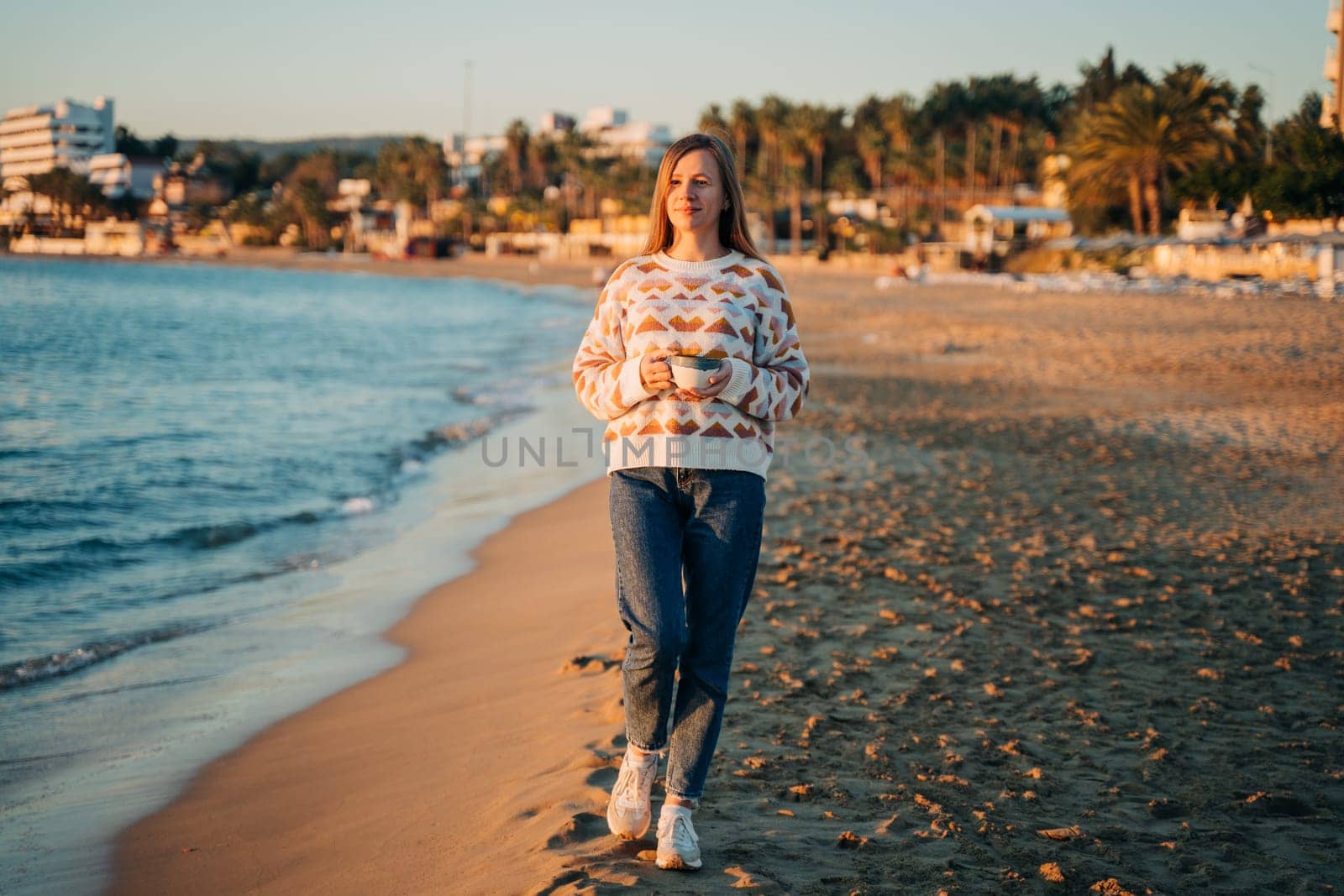 Young beautiful girl in cozy sweater holding coffee cup while enjoying winter sun on seaside shore during mild sunset. Cute attractive woman enjoying her cup of tea while watching autumn ocean waves by Ostanina