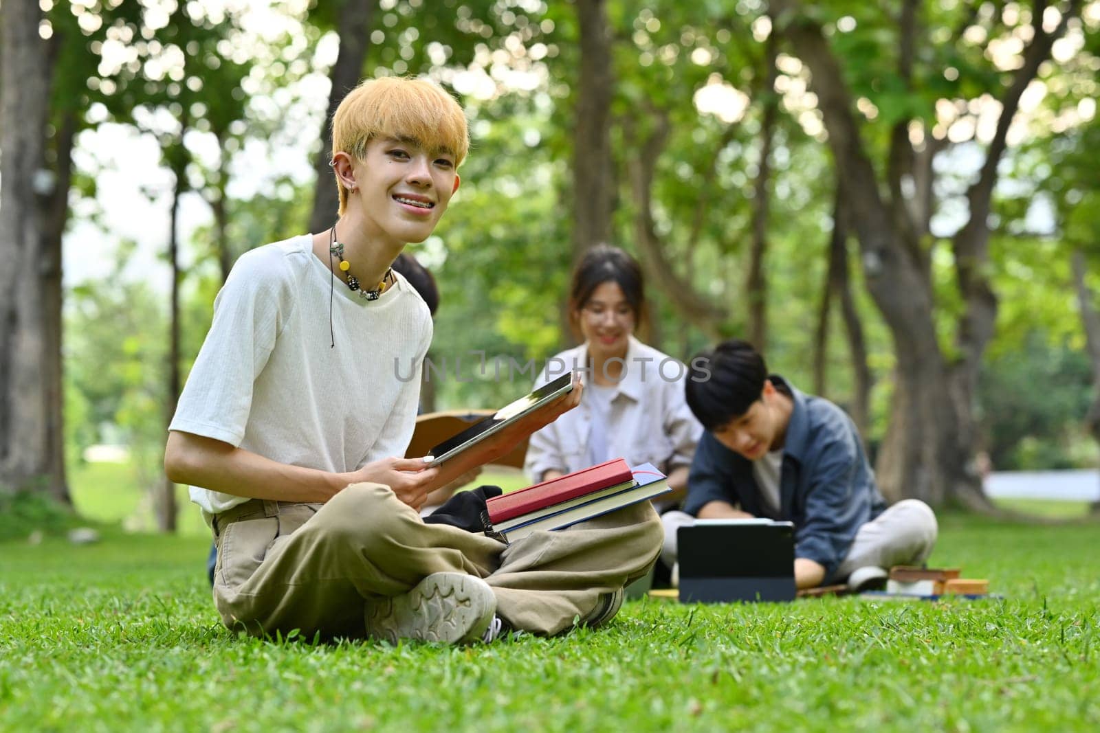 Asian male student sitting on green grass at the college campus on a sunny day. Education and lifestyle concept.
