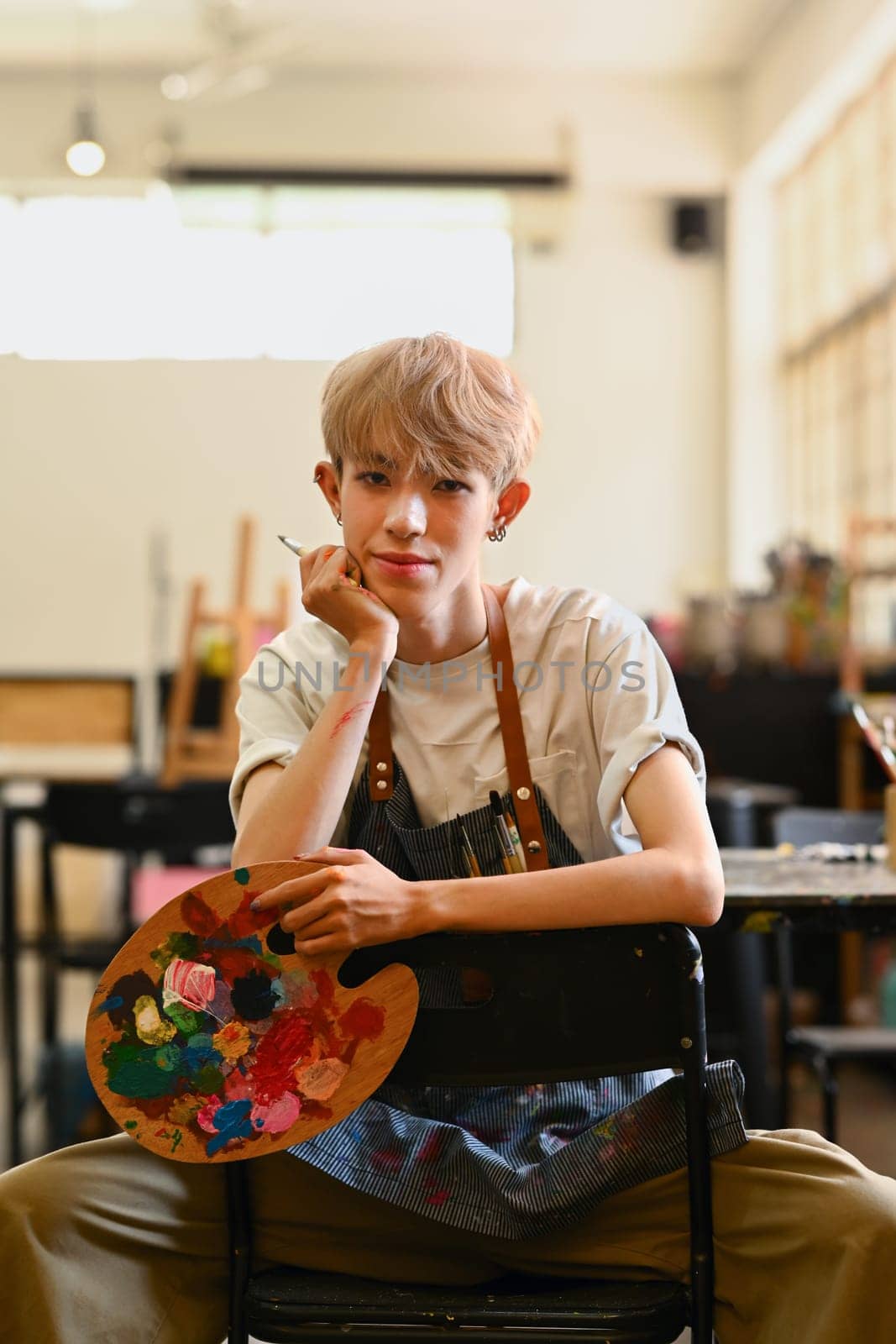 Portrait of young hipster male artist wearing apron holding palette and brush posing in art studio by prathanchorruangsak