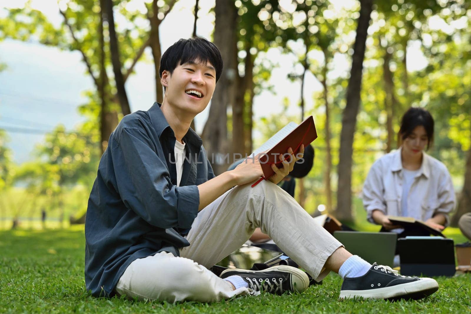 Smiling asian student man sitting on the grass and reading a book. Youth lifestyle and education.
