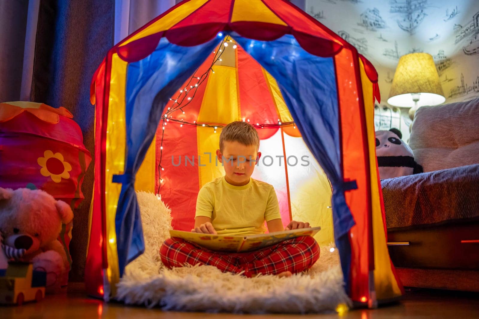 Cute little boy is reading a book in the evening in the tent.