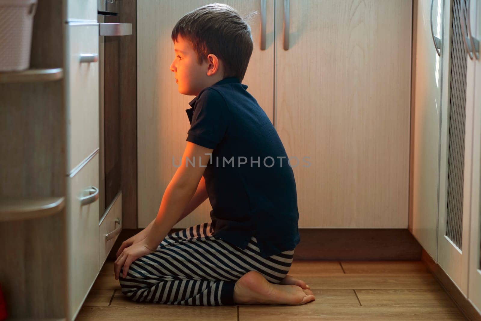 A child is sitting near the oven in the kitchen and waiting. Curious boy is watching through the glass of kitchen oven. Baking pizza, muffins , cupcakes or cookies.