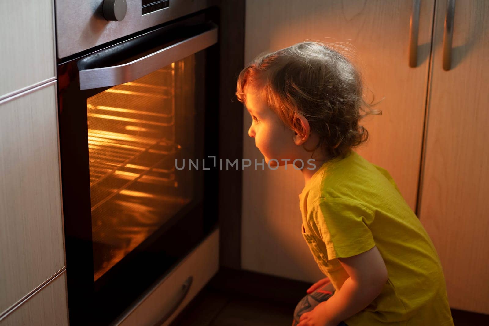 A child is sitting near the oven in the kitchen and waiting. Curious boy is watching through the glass of kitchen oven. Baking pizza, muffins , cupcakes or cookies.