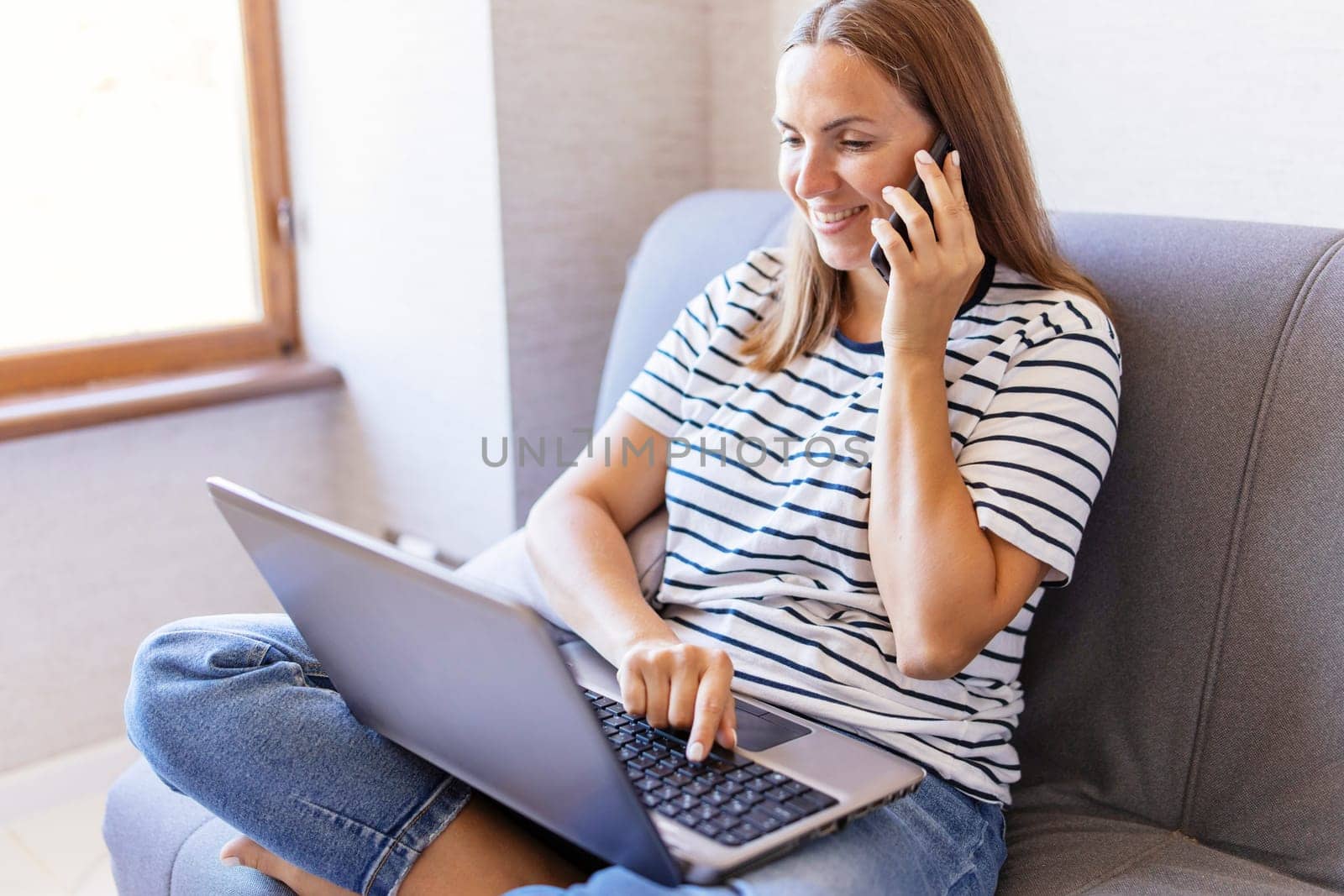Happy young woman using mobile phone while sitting on sofa at home with laptop. A woman is talking on a cell phone, asks questions, looks at the computer screen.