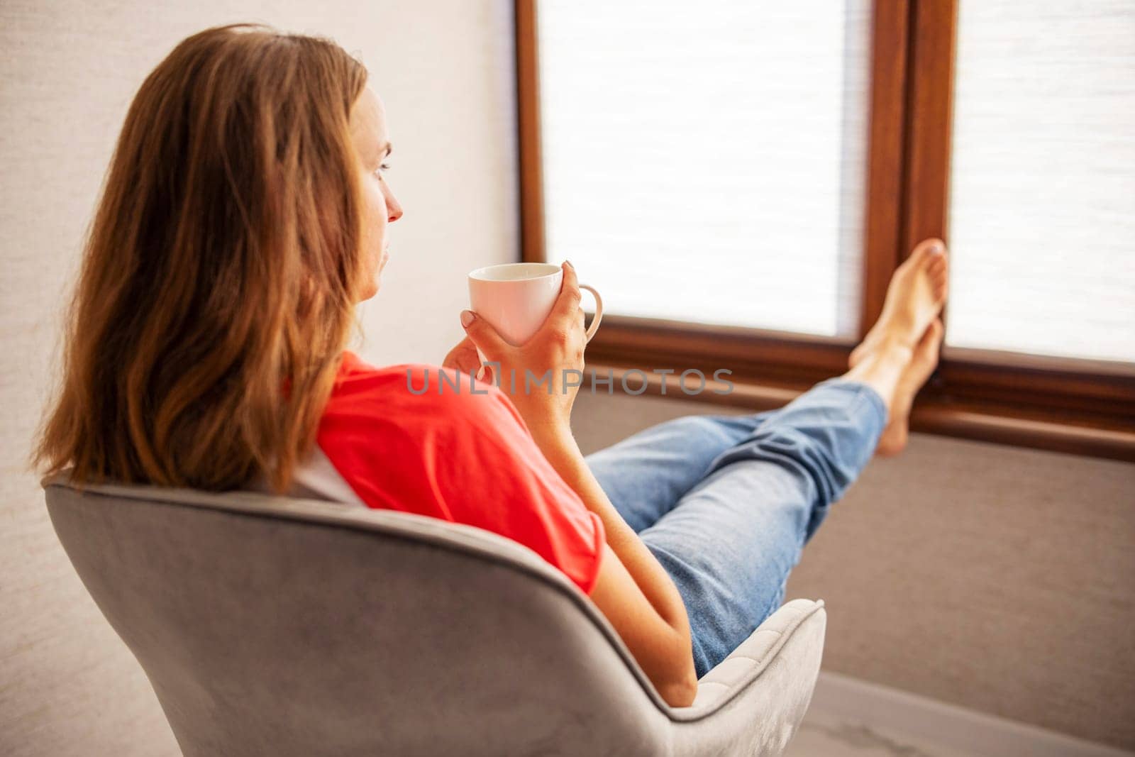 Young beautiful woman in casual wear sitting in armchair with cup of hot coffee. Happy woman relaxing in armchair near a window.