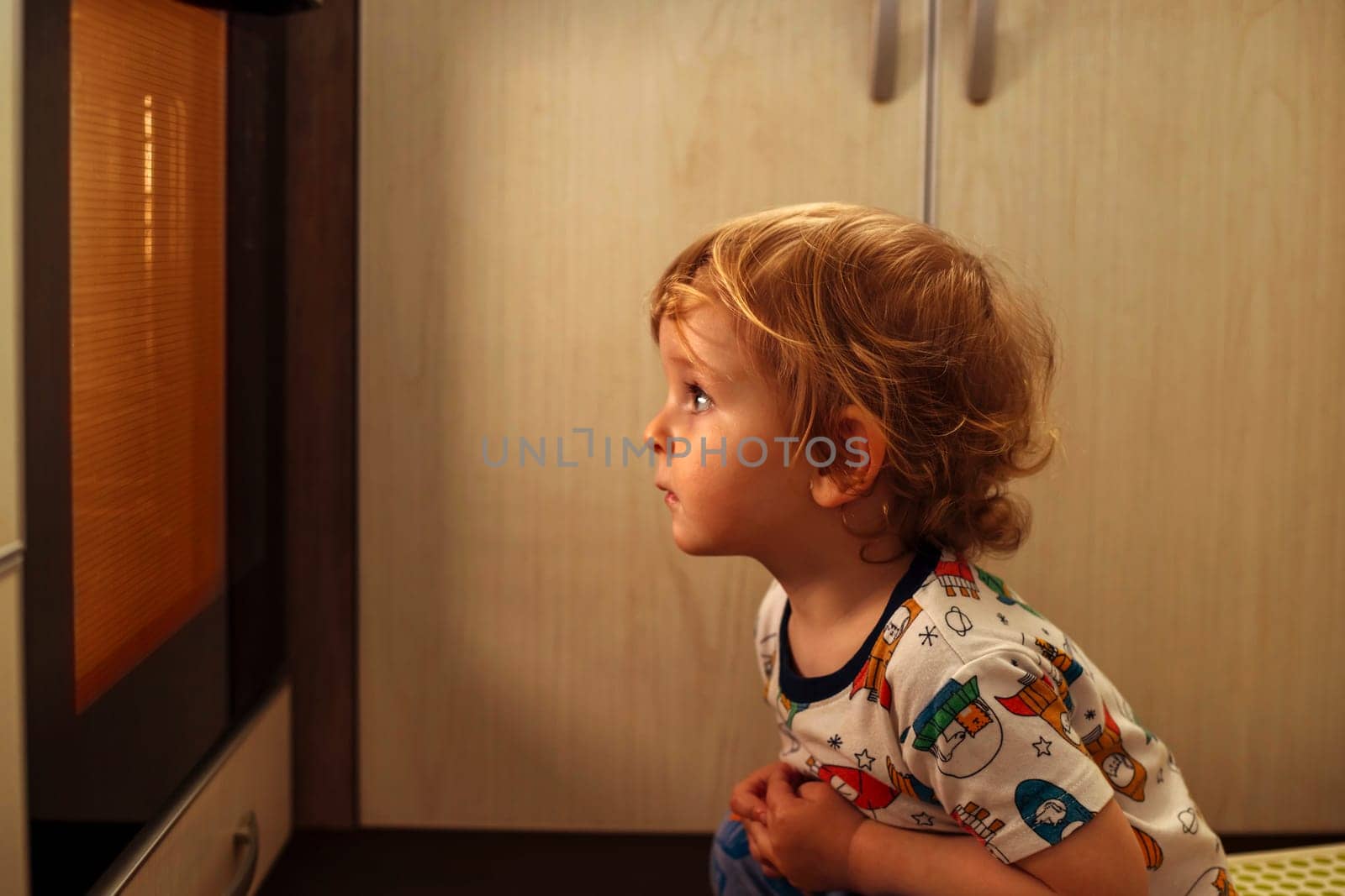 A child is sitting near the oven in the kitchen and waiting. Curious boy is watching through the glass of kitchen oven. Baking pizza, muffins , cupcakes or cookies.