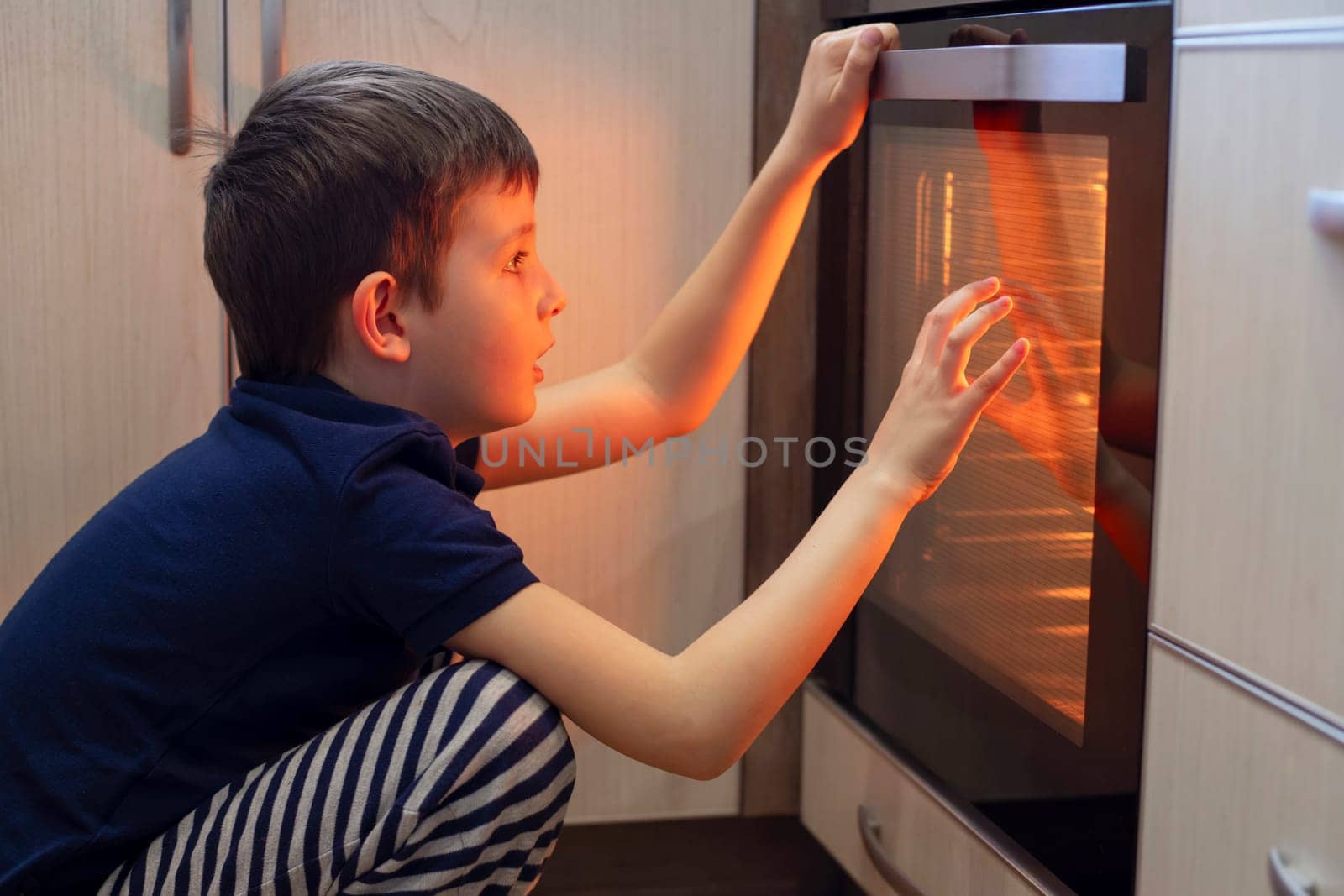 A child is sitting near the oven in the kitchen and waiting. Curious boy is watching through the glass of kitchen oven. Baking pizza, muffins, cupcakes or cookies.