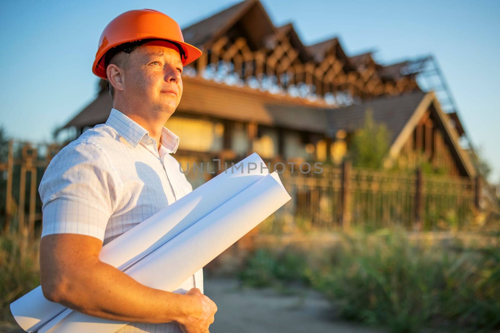 Young engineer in a yellow helmet with blueprints at a construction site in front of a building requiring reconstruction or repair.