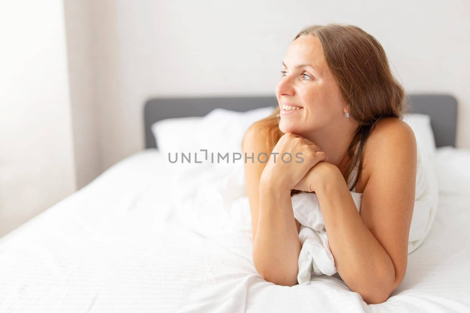 Young smiling attractive brunette woman lies in a big white bed.