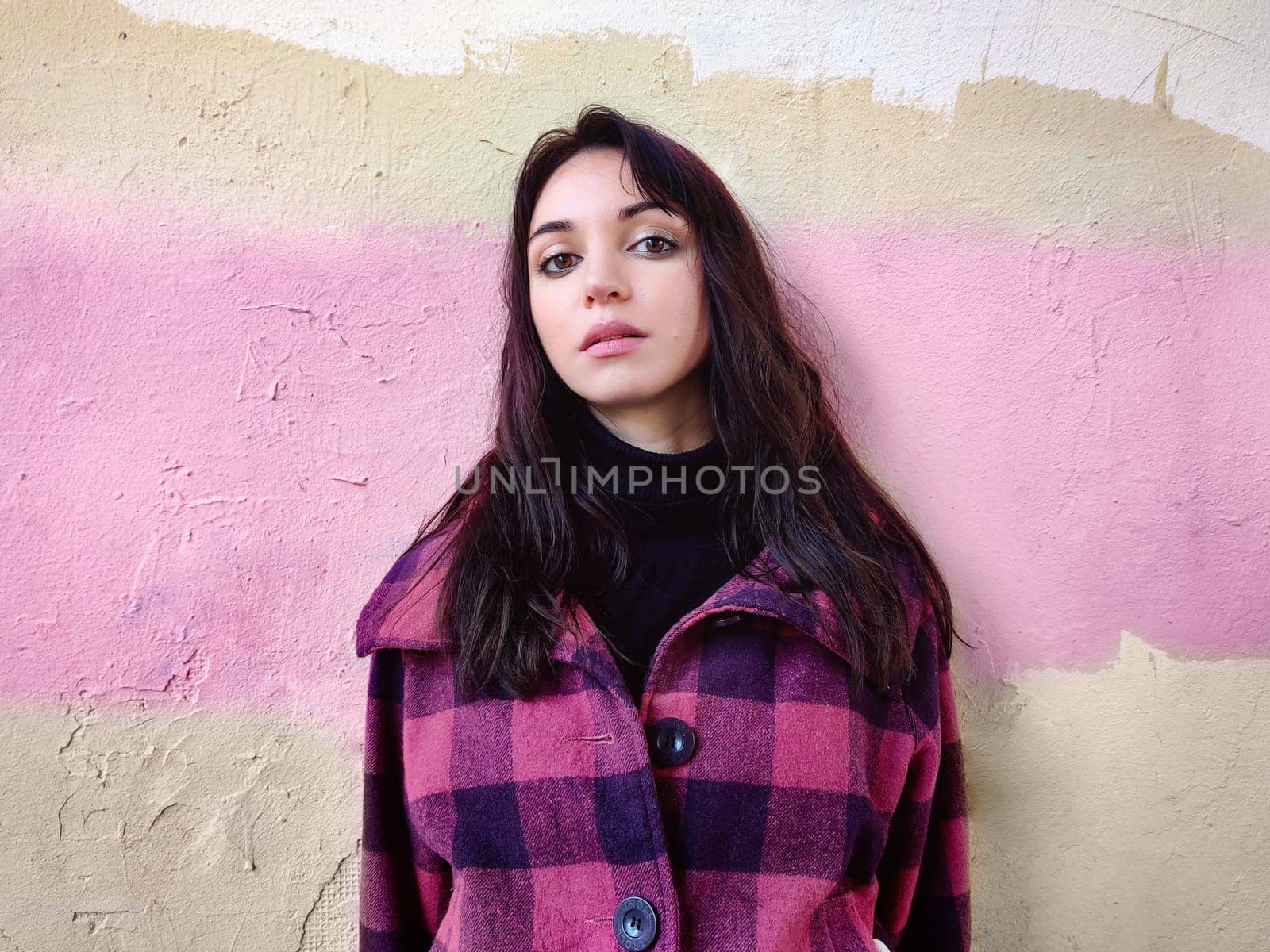 Thoughtful woman with dark hair, in black golf and in a purple plaid coat against the background of a colored wall. Thoughtful state.