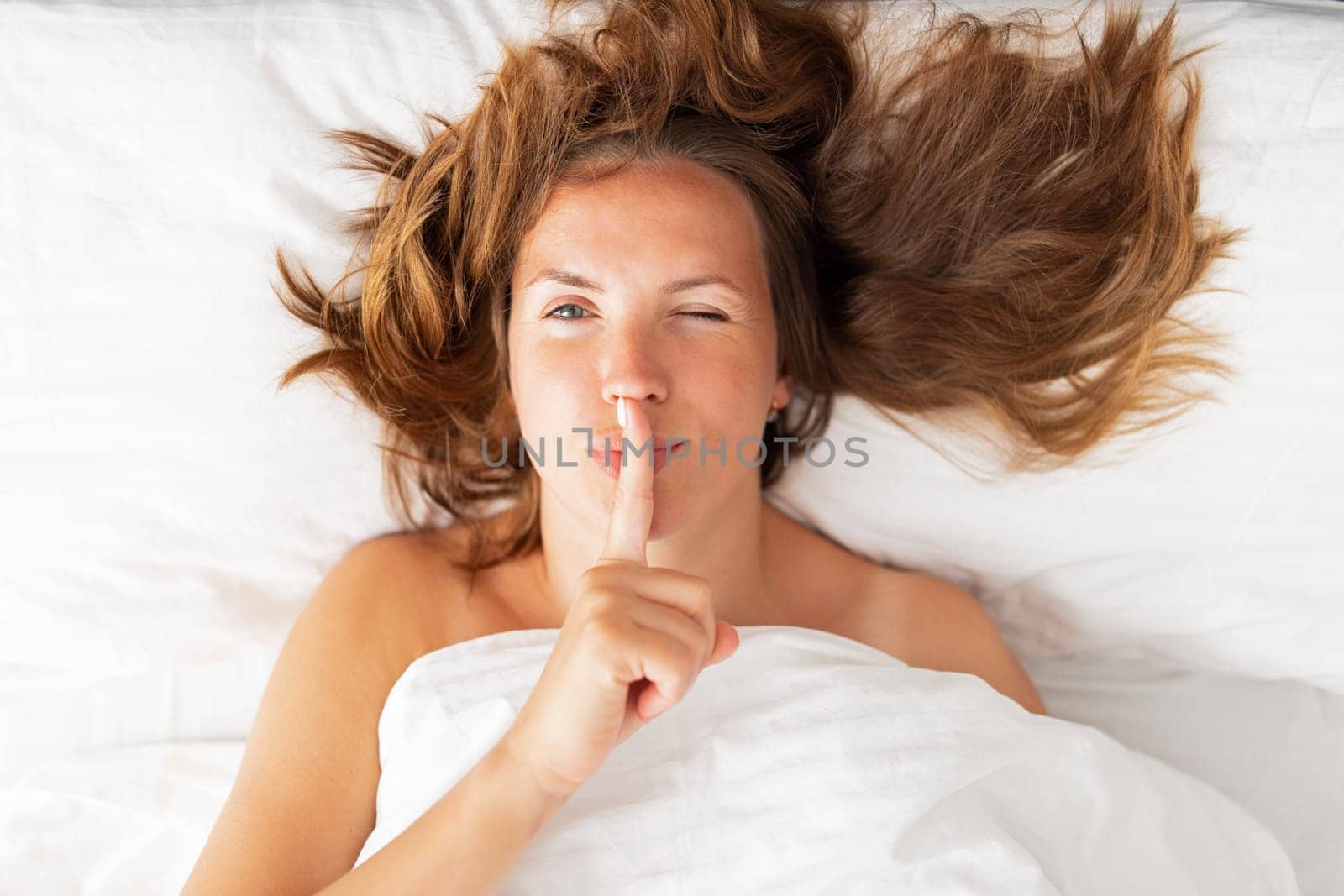 Women's secrets concept. Portrait of a woman lying in bed covered with a blanket holding finger on lips, asking to be quiet, gesturing and showing shh hush sign.