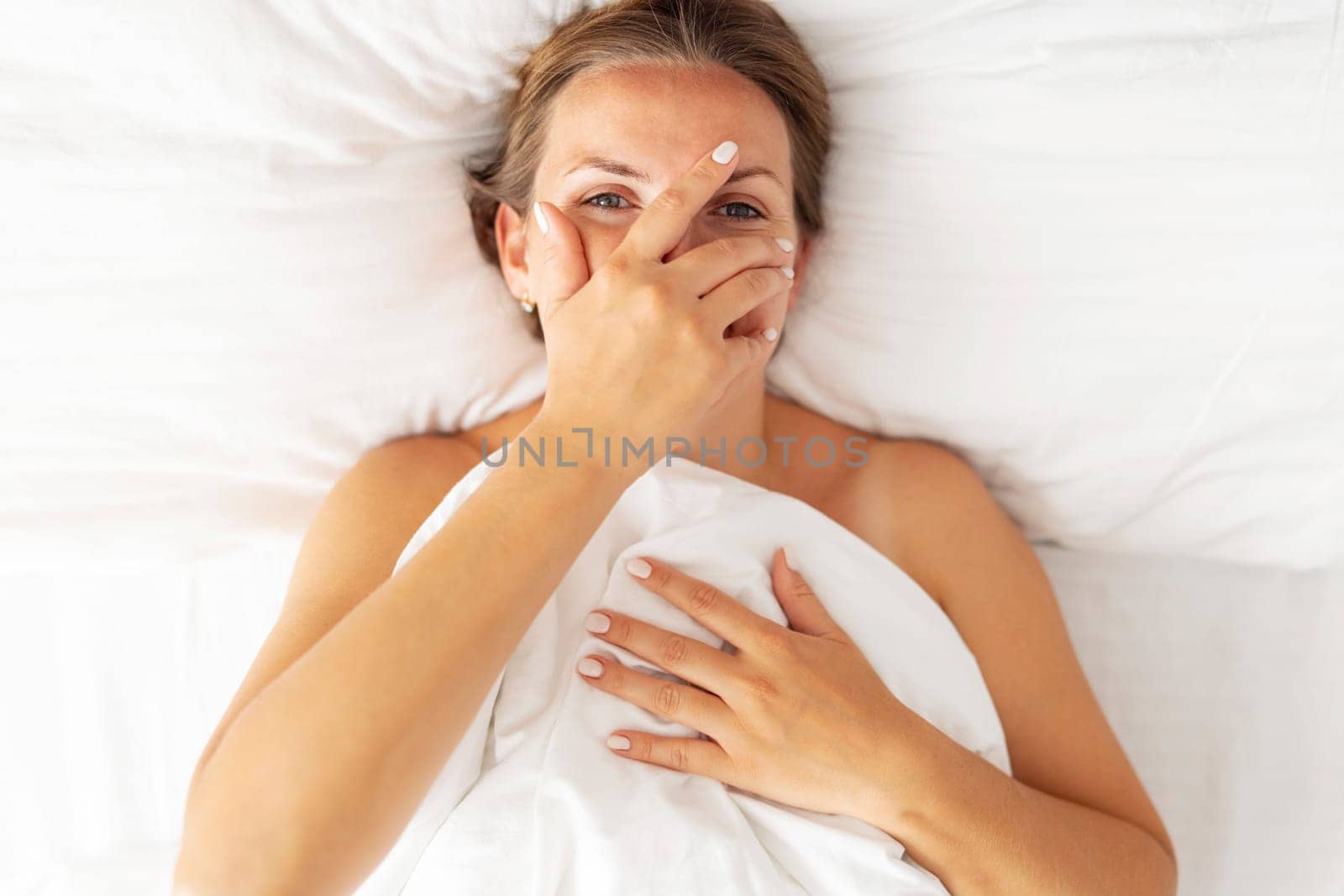 Top view of happy young woman peeking through her fingers and smiling while lying in bed.