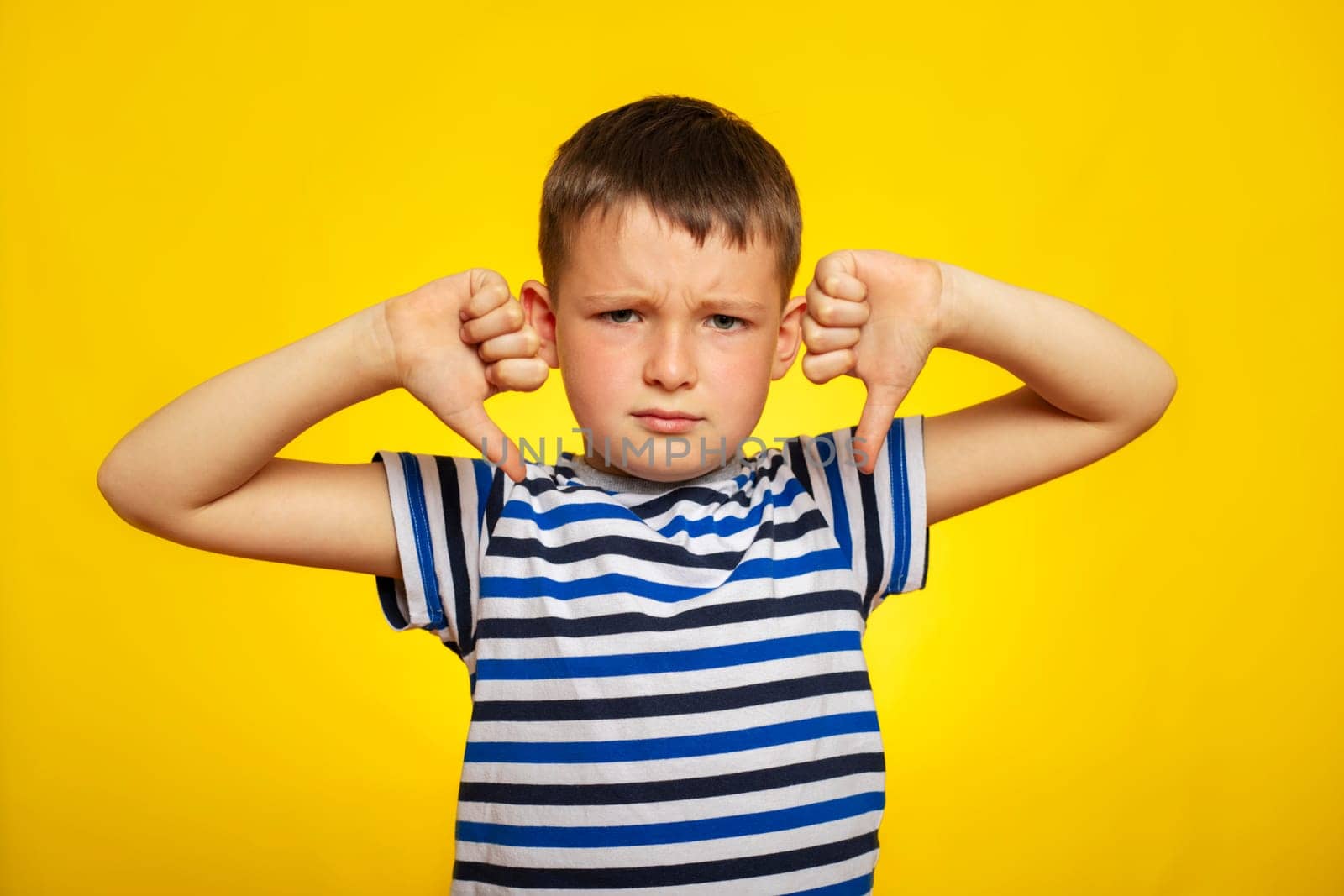 A young boy in striped t-shirt showing thumbs down gesture with two hands on yellow background. Displeased child giving thumbs down hand gesture.