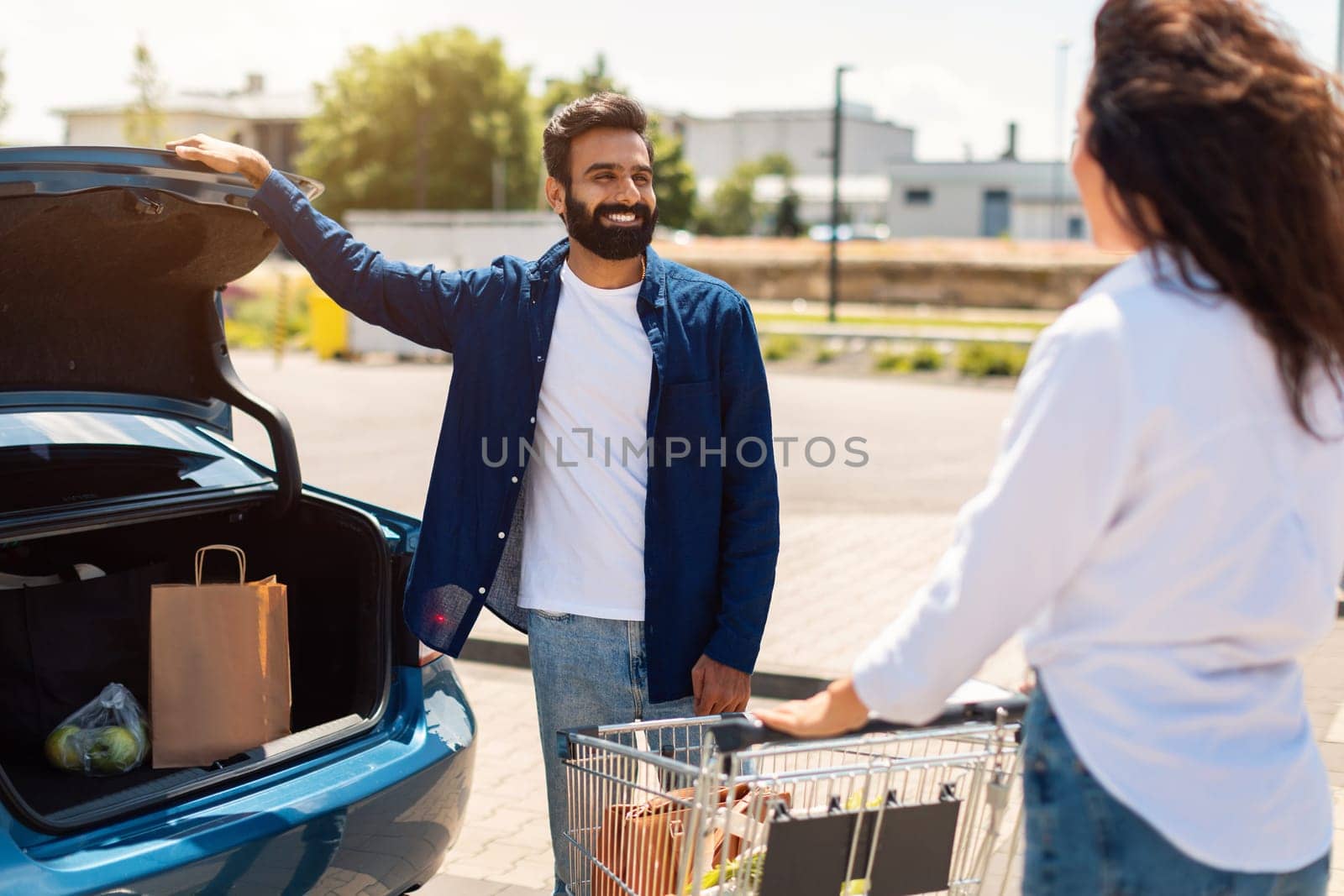 Happy arab couple with shopping cart full of fresh food, packing products into the car on the outdoor parking, woman standing near shopping cart