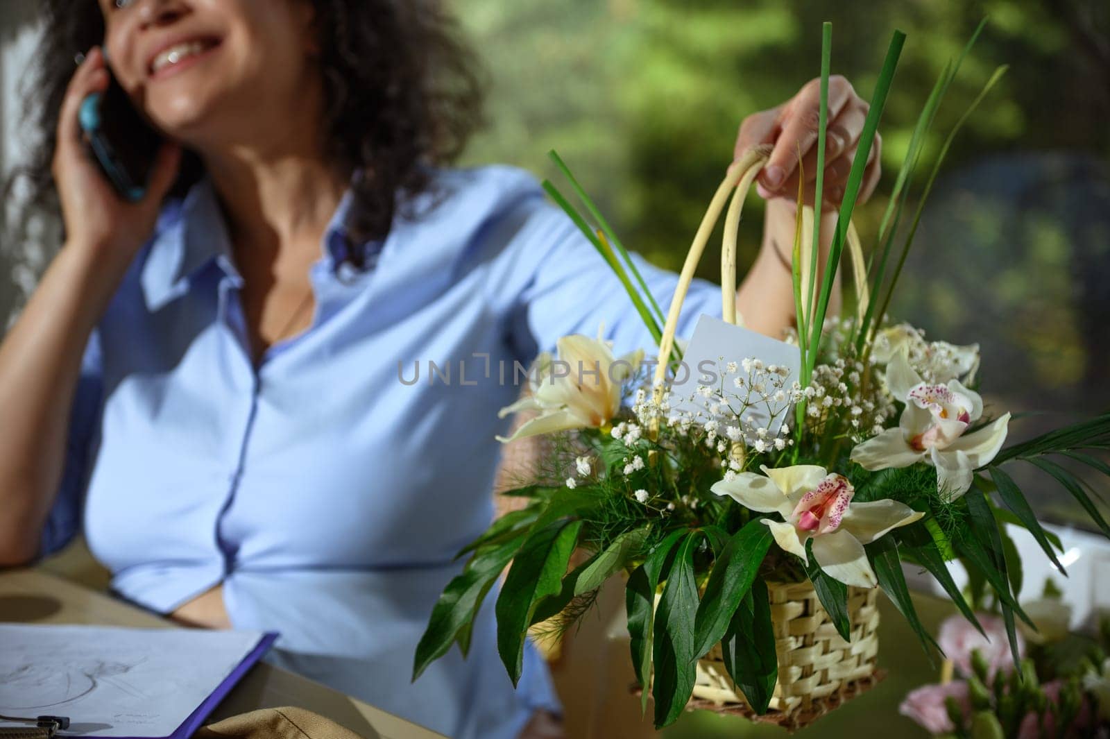 Selective focus on a beautiful flower arrangement with orchids and fresh plants in wicker basket over blurred female florist discussing delivery the order on mobile phone, in floristry design studio