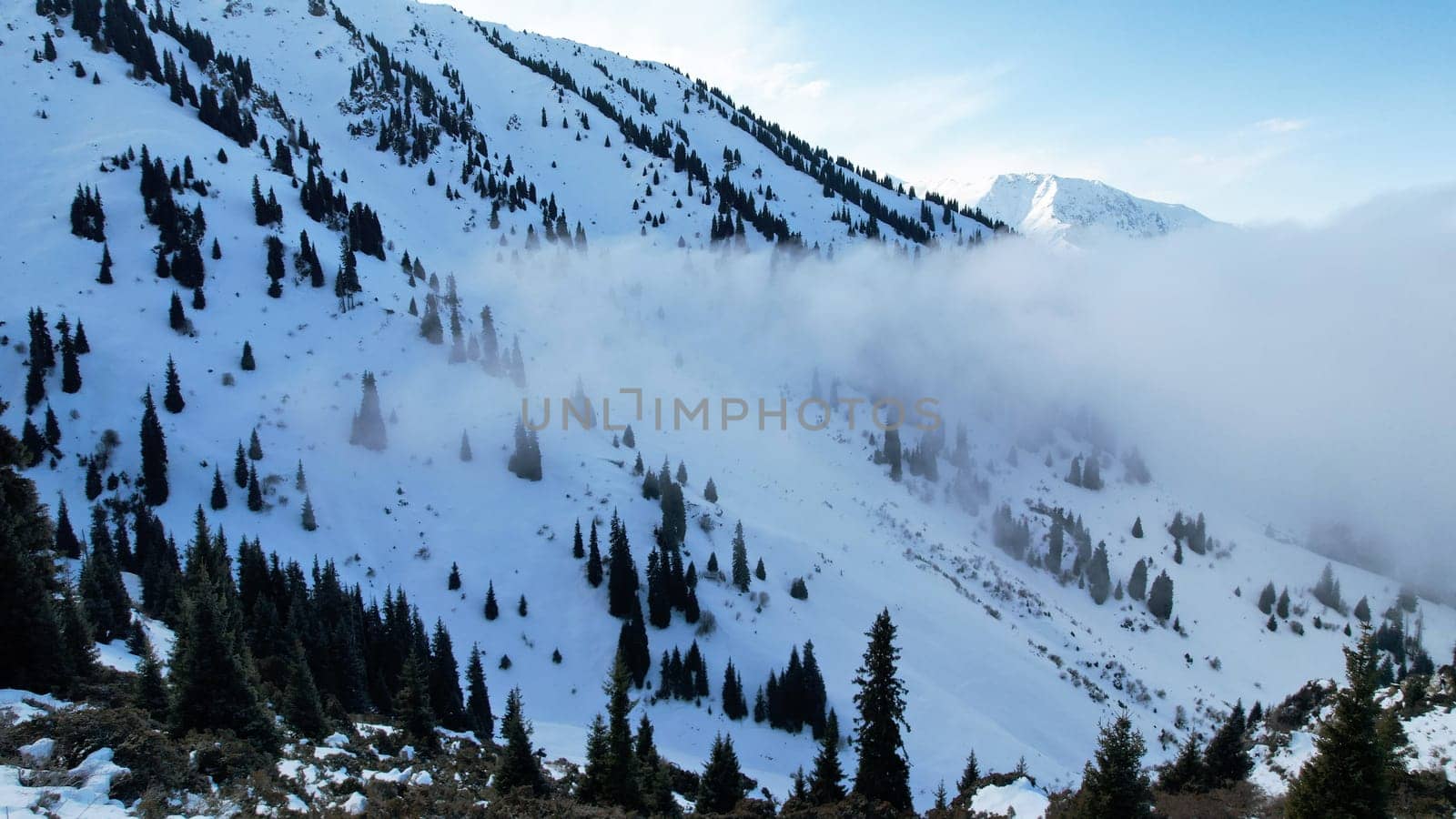 White cumulus clouds in snowy mountains in winter. The rays of the sun fall on part of the clouds, a shadow comes from the peak. Blue sky. Coniferous fir trees grow on the hills. Tourists are walking