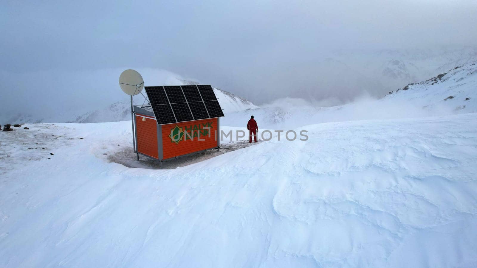A rescue hut high in the mountains among the clouds. There is a climber standing next to the house. White clouds enveloped the snowy mountains. Yellow dawn and clouds. Snowy hills. 01.05.2023 Almaty