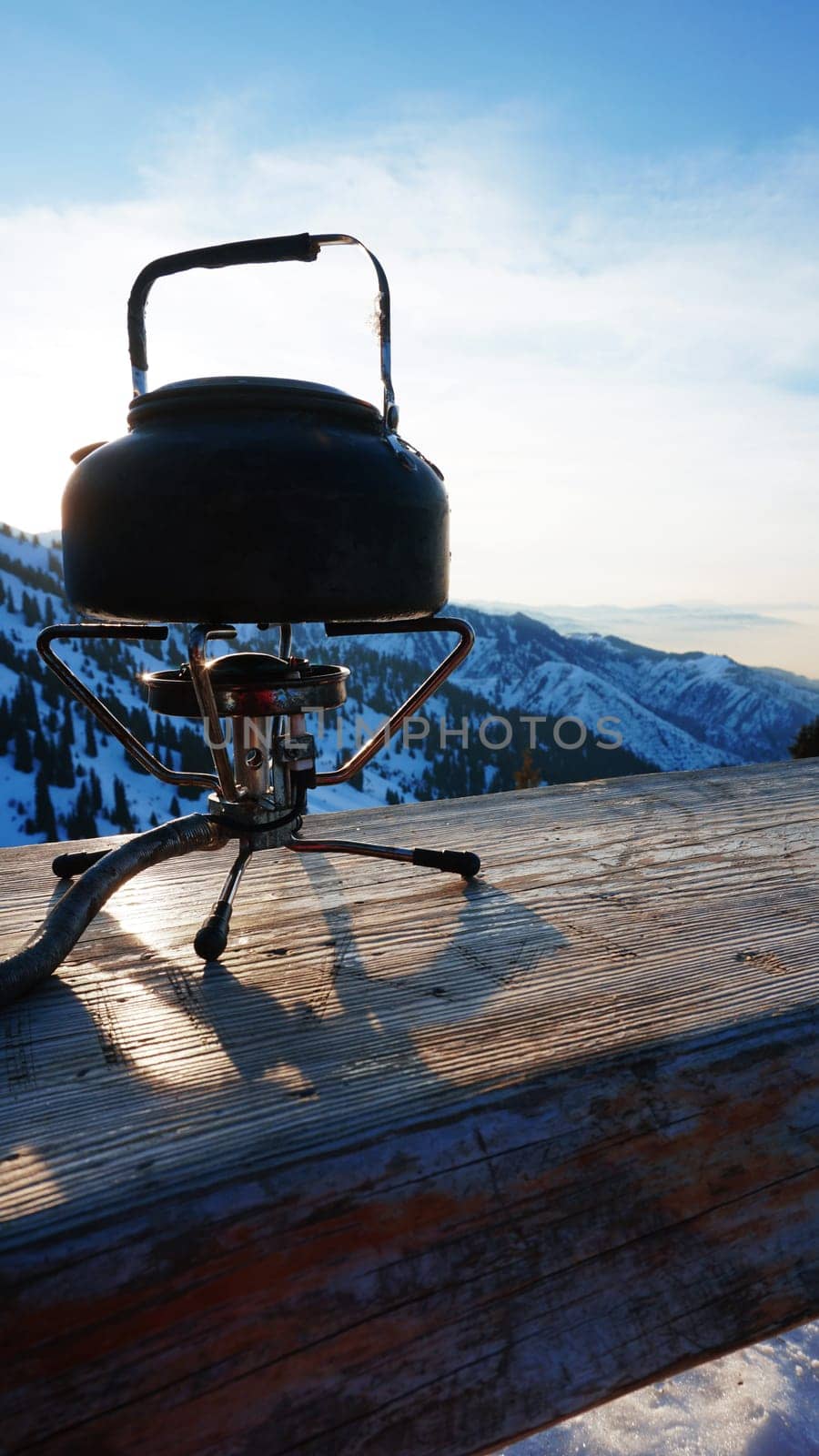 The burner is burning with a blue fire with a kettle. The burner and the cylinder are on the bench. In the background there are green fir trees, mountains in white snow and a beautiful sunset