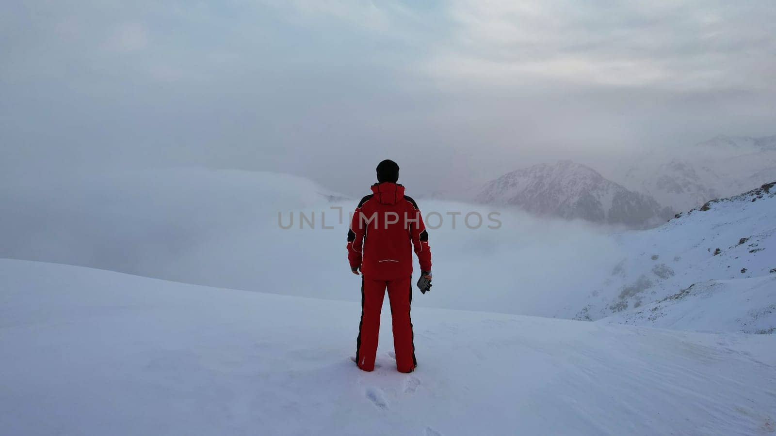 A climber in a red suit stands on top of mountain by Passcal