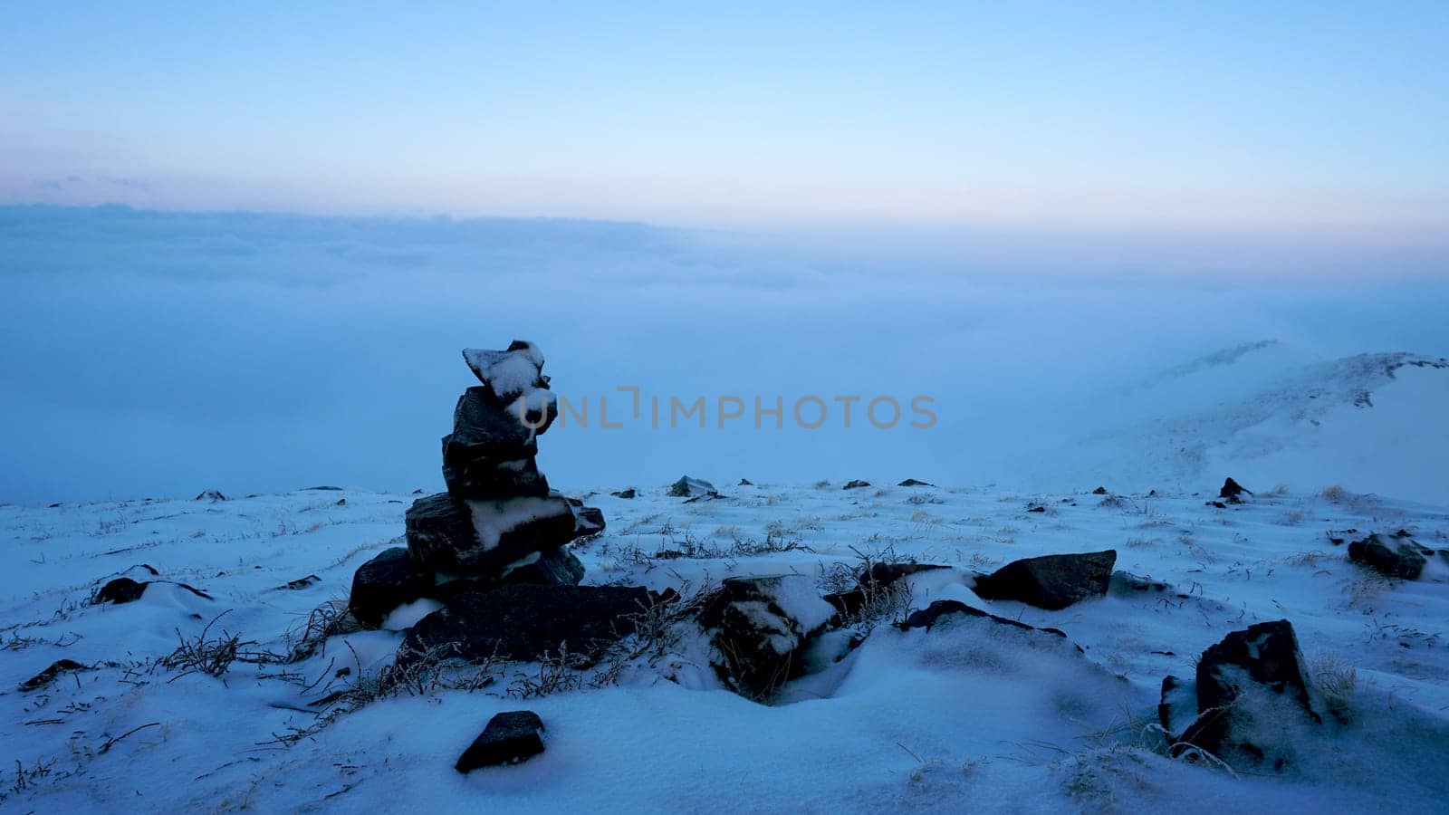 An ocean of clouds in the snowy mountains at dawn. White clouds are like a carpet in a gorge. Waves rise and descend from the mountains. Sunrise of the yellow sun. There are signposting stones. Almaty