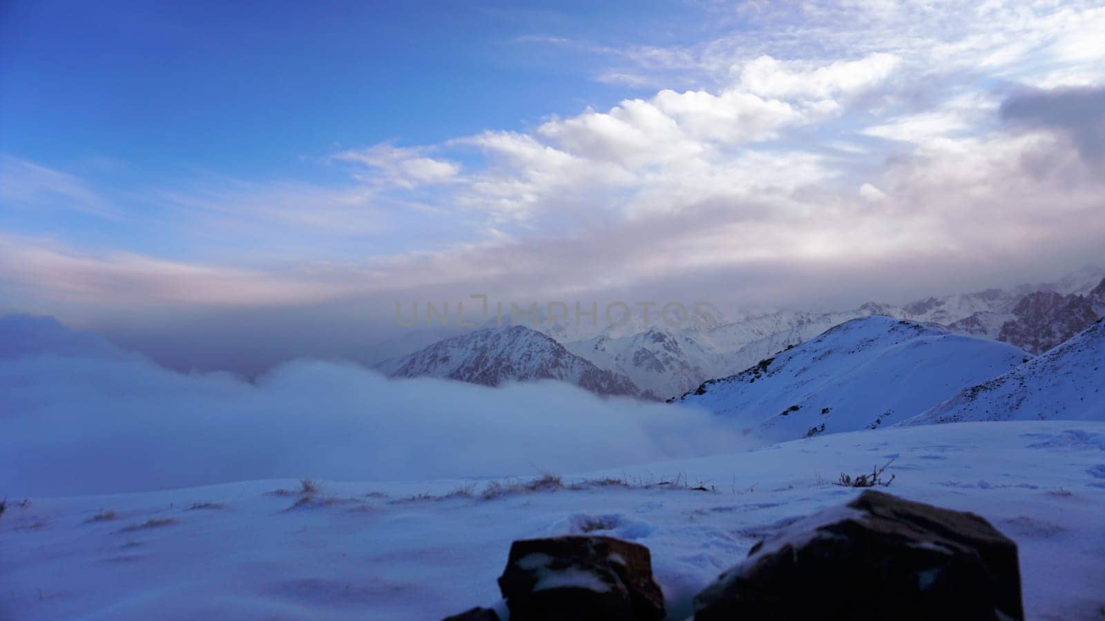 An ocean of clouds in the snowy mountains at dawn. White clouds are like a carpet in a gorge. Waves rise and descend from the mountains. Sunrise of the yellow sun. There are stones lying. Kazakhstan