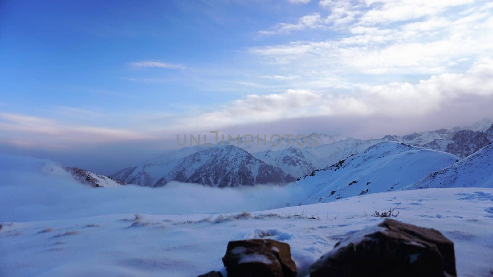 An ocean of clouds in the snowy mountains at dawn. White clouds are like a carpet in a gorge. Waves rise and descend from the mountains. Sunrise of the yellow sun. There are stones lying. Kazakhstan