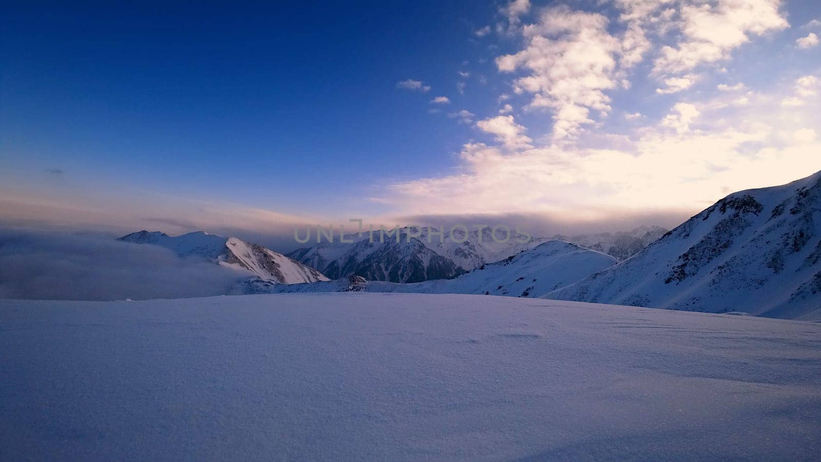 An ocean of clouds in the snowy mountains at dawn. White clouds are like a carpet in a gorge. Waves rise and descend from the mountains. Sunrise of the yellow sun. Clear sky, mountain peaks. Almaty