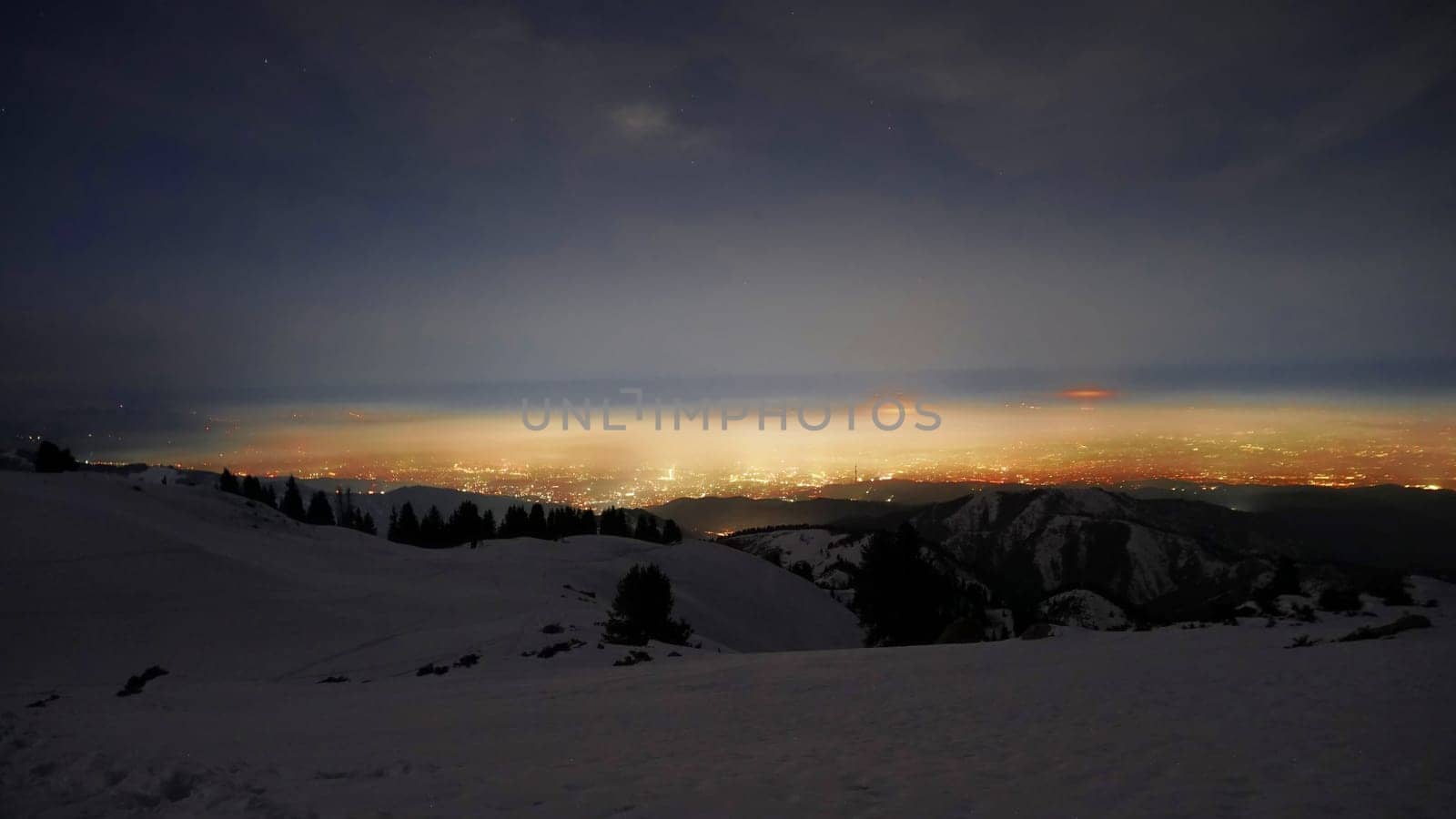 View of the night city from the snowy mountains. The glow from the city rises to the clouds. Bright lights are visible. In places, even the stars are visible. Steep cliffs, big rocks and snow. Almaty