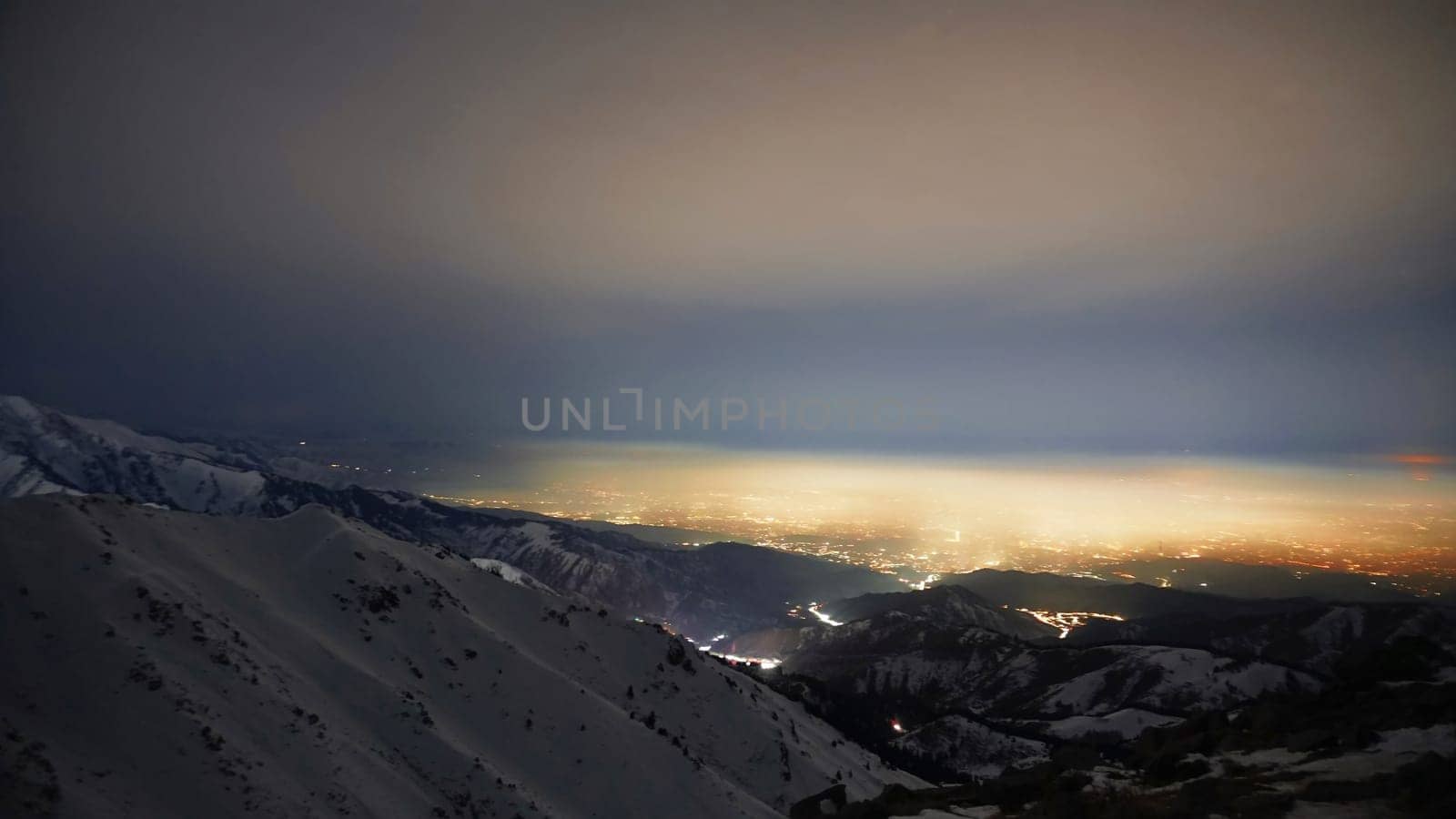 View of the night city from the snowy mountains. The glow from the city rises to the clouds. Bright lights are visible. In places, even the stars are visible. Steep cliffs, big rocks and snow. Almaty