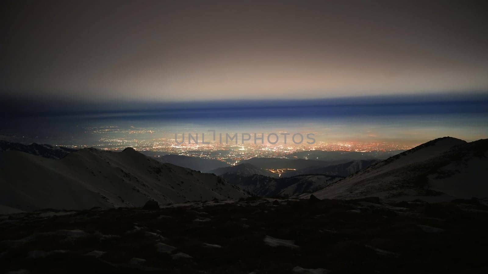 View of the night city from the snowy mountains. The glow from the city rises to the clouds. Bright lights are visible. In places, even the stars are visible. Steep cliffs, big rocks and snow. Almaty