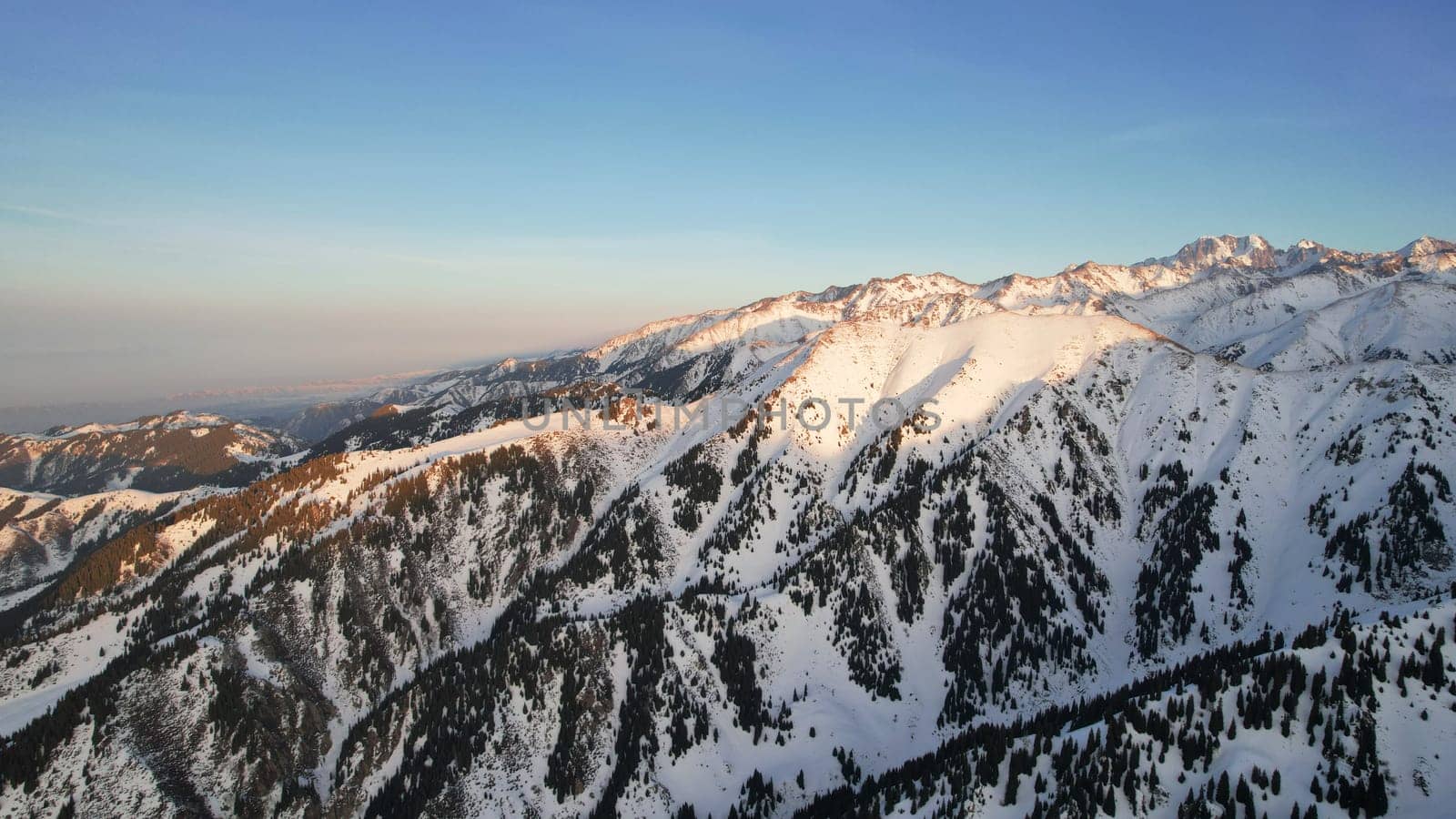 Epic red-orange sunset in high snowy mountains. There are tall coniferous trees, there is snow on the hills. High peaks. The sun's rays fall on the clouds and fog that is lower down the gorge. Almaty