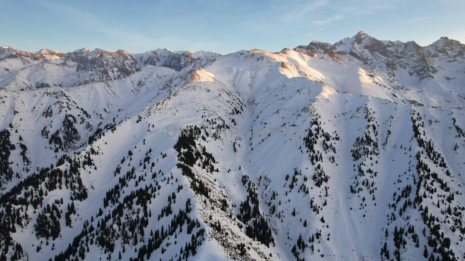 Epic red-orange sunset in high snowy mountains. There are tall coniferous trees, there is snow on the hills. High peaks. The sun's rays fall on the clouds and fog that is lower down the gorge. Almaty