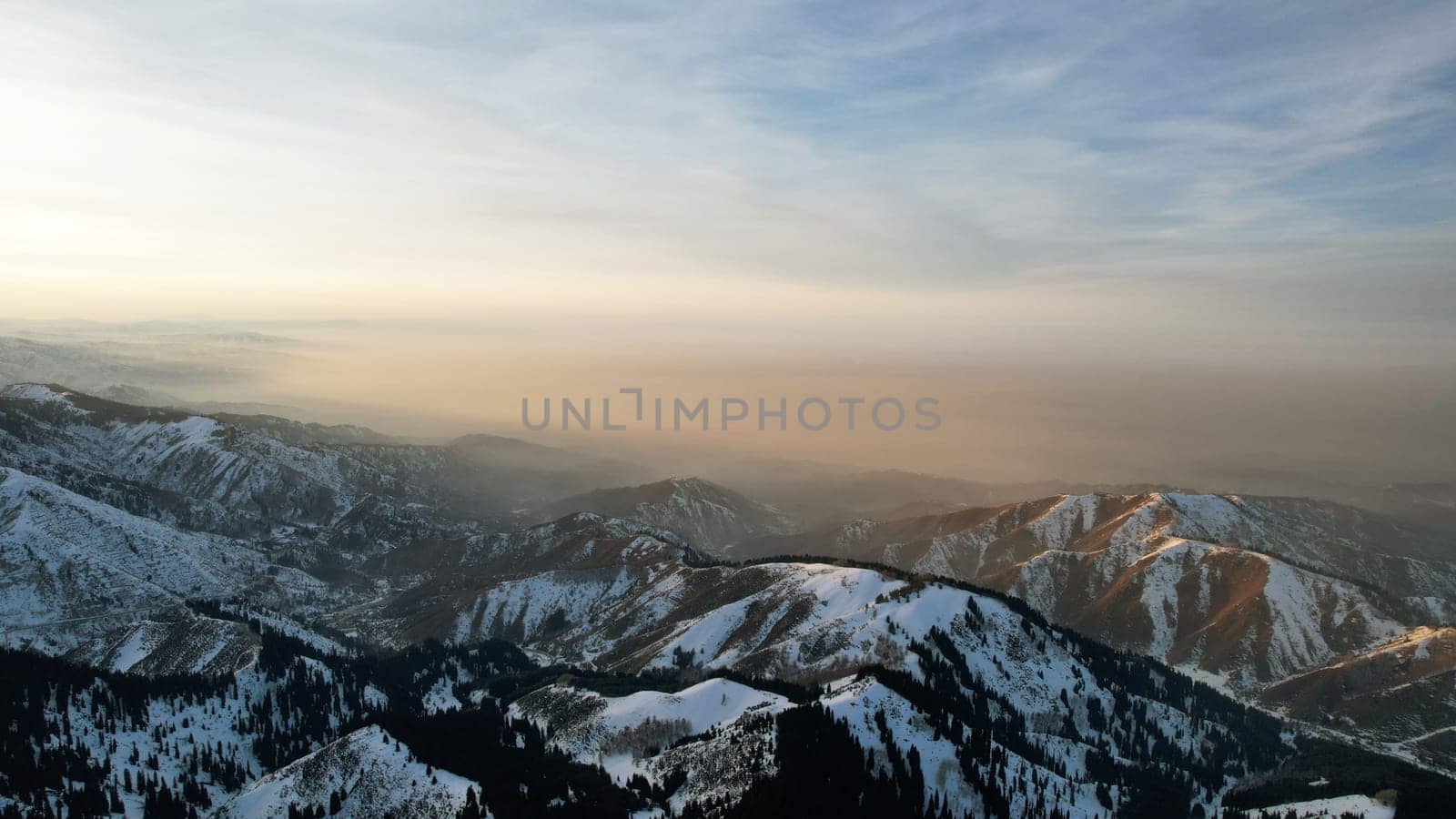 Epic red-orange sunset in high snowy mountains. There are tall coniferous trees, there is snow on the hills. High peaks. The sun's rays fall on the clouds and fog that is lower down the gorge. Almaty