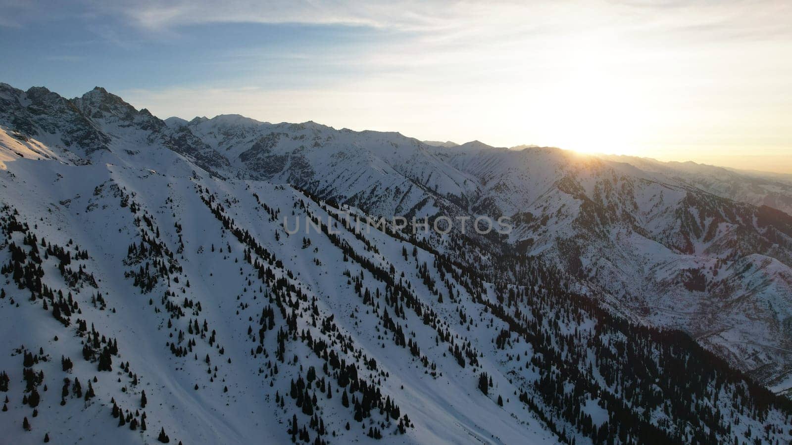 Epic red-orange sunset in high snowy mountains. There are tall coniferous trees, there is snow on the hills. High peaks. The sun's rays fall on the clouds and fog that is lower down the gorge. Almaty