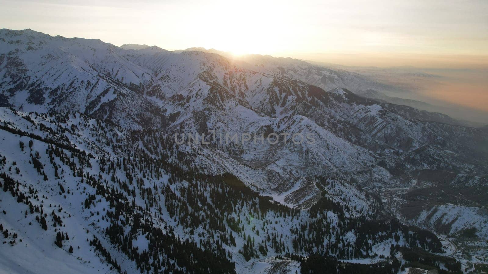 Epic red-orange sunset in high snowy mountains. There are tall coniferous trees, there is snow on the hills. High peaks. The sun's rays fall on the clouds and fog that is lower down the gorge. Almaty