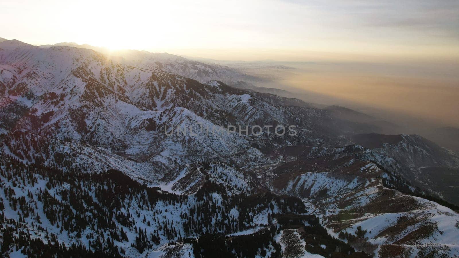 Epic red-orange sunset in high snowy mountains. There are tall coniferous trees, there is snow on the hills. High peaks. The sun's rays fall on the clouds and fog that is lower down the gorge. Almaty