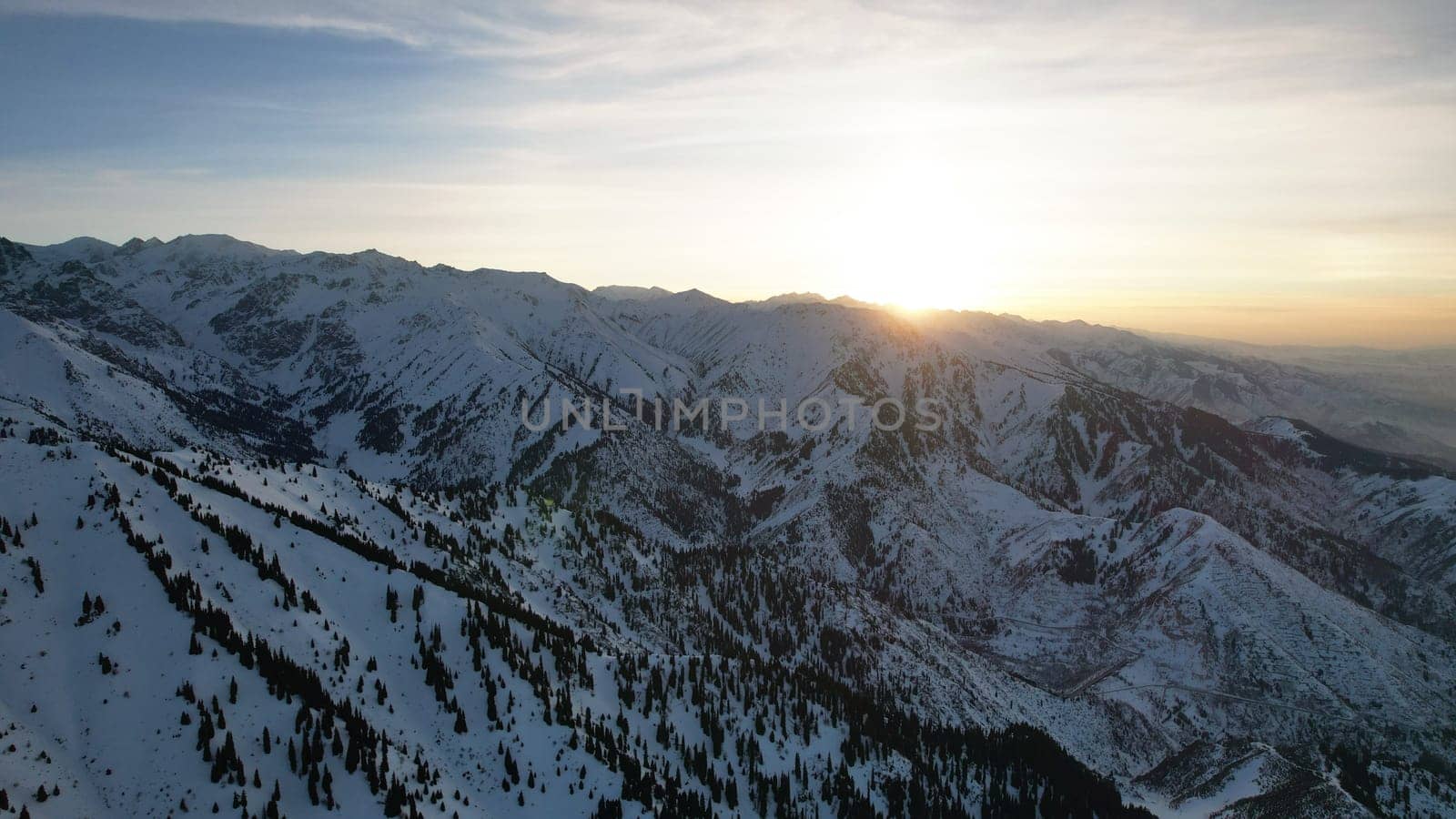 Epic red-orange sunset in high snowy mountains. There are tall coniferous trees, there is snow on the hills. High peaks. The sun's rays fall on the clouds and fog that is lower down the gorge. Almaty