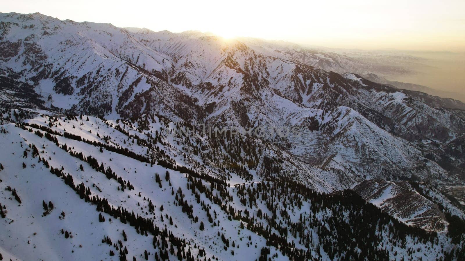 Epic red-orange sunset in high snowy mountains. There are tall coniferous trees, there is snow on the hills. High peaks. The sun's rays fall on the clouds and fog that is lower down the gorge. Almaty