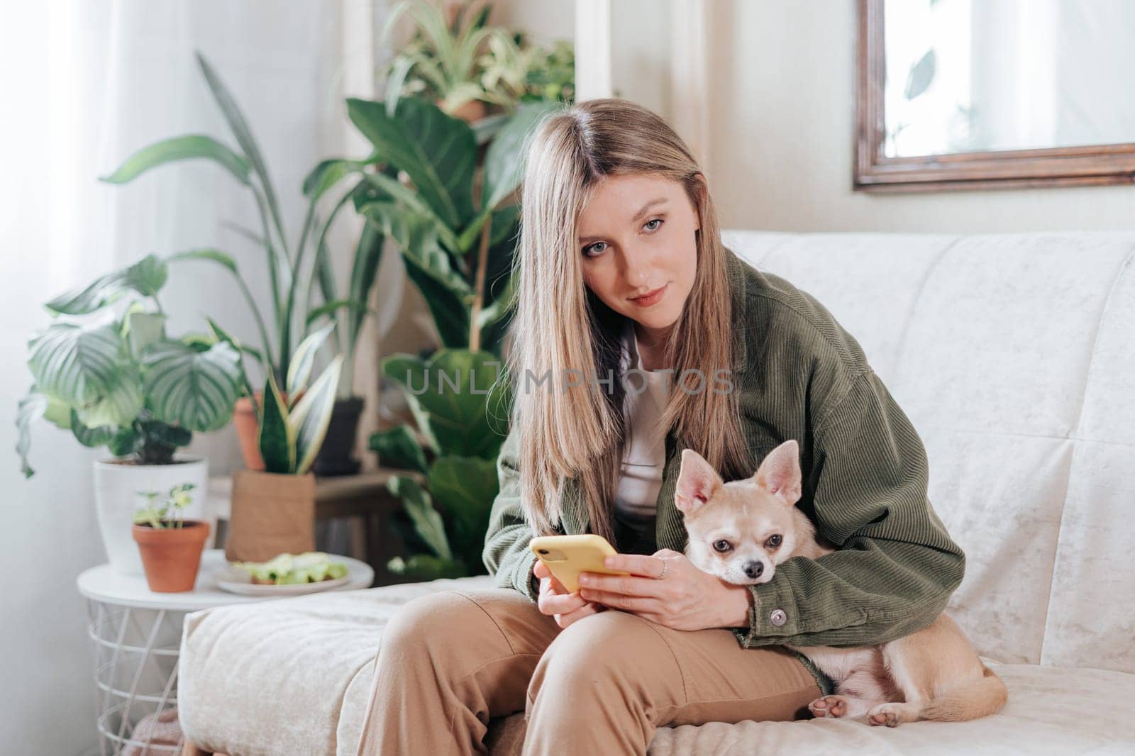 Freelance woman with mobile phone and dog, and working from home office. Happy girl sitting on couch in living room with plants. Distance learning and online education by Ostanina