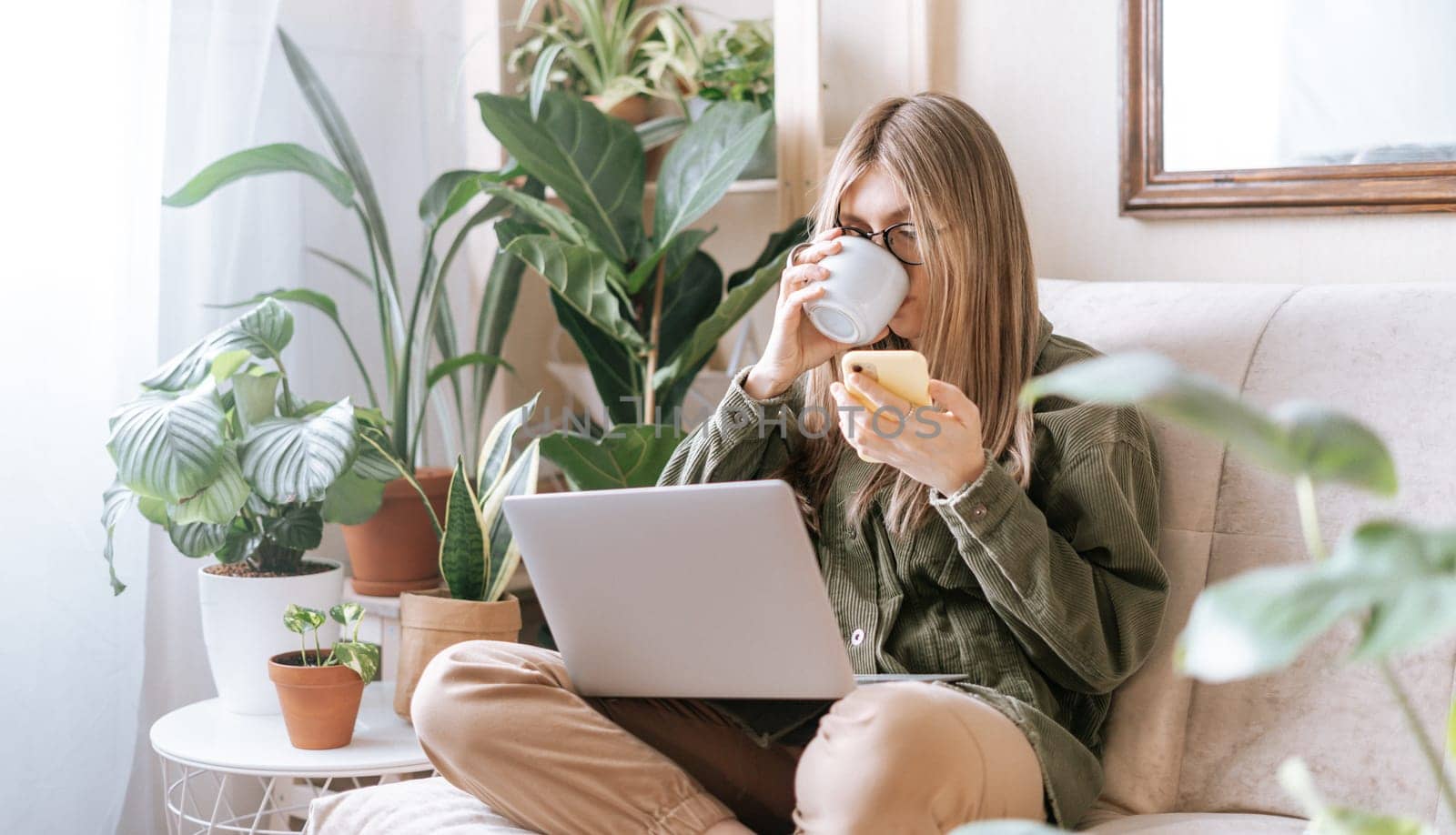 Freelance woman in glasses with mobile phone typing at laptop and working from home office. Happy girl sitting on couch in living room with plants. Distance learning online education and work.