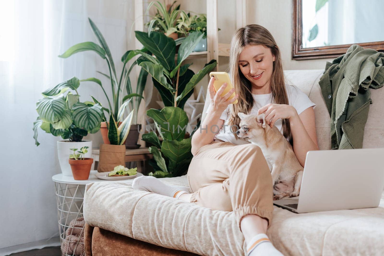 Freelance woman with dog and laptop, texting at mobile phone and working from home office. Happy girl sitting on couch in living room with plants. Distance learning online education and work by Ostanina