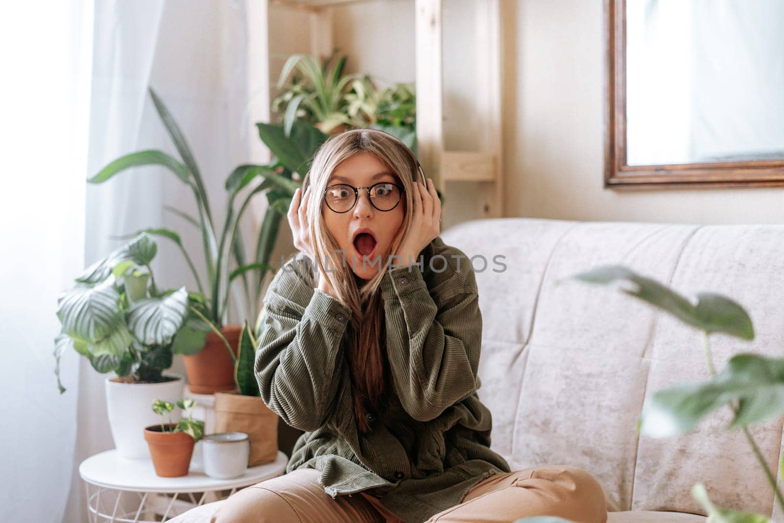 Shocked woman with open mouth in glasses listening music in headphones and relax at home. Frustrated girl sitting on couch in living room with plants. Urban jungle concept