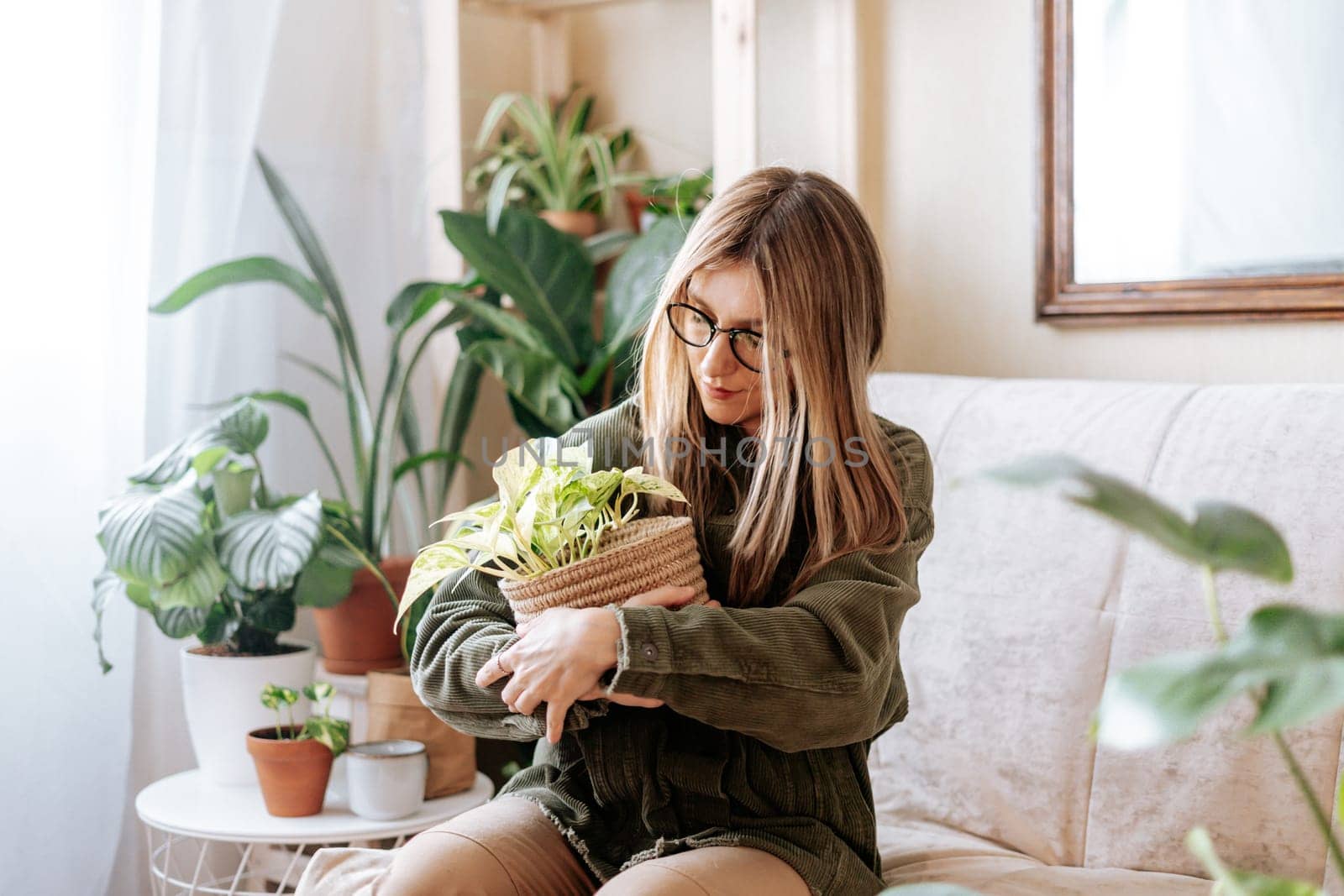 Freelance woman gardener in glasses holding and hug wicker basket with plant and working from home office. Happy girl sitting relaxing on couch in living room with plants in cozy home. Urban jungle.
