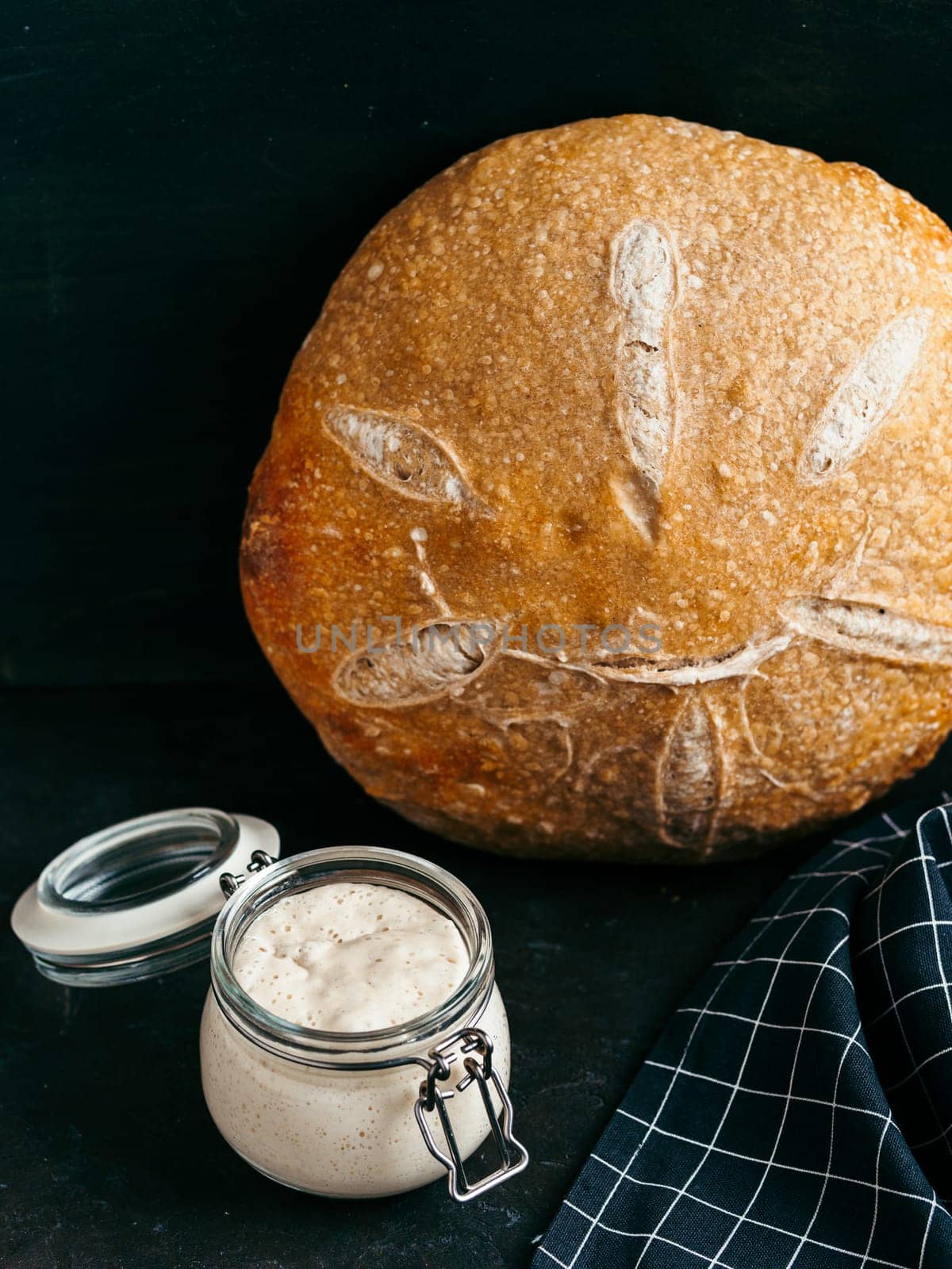 Sourdoug starter and wheat sourdough bread. Wheat sour dough starter in glass jar and delicious homemade round sourdough bread on black background. Home made sourdough bread making concept. Vertical