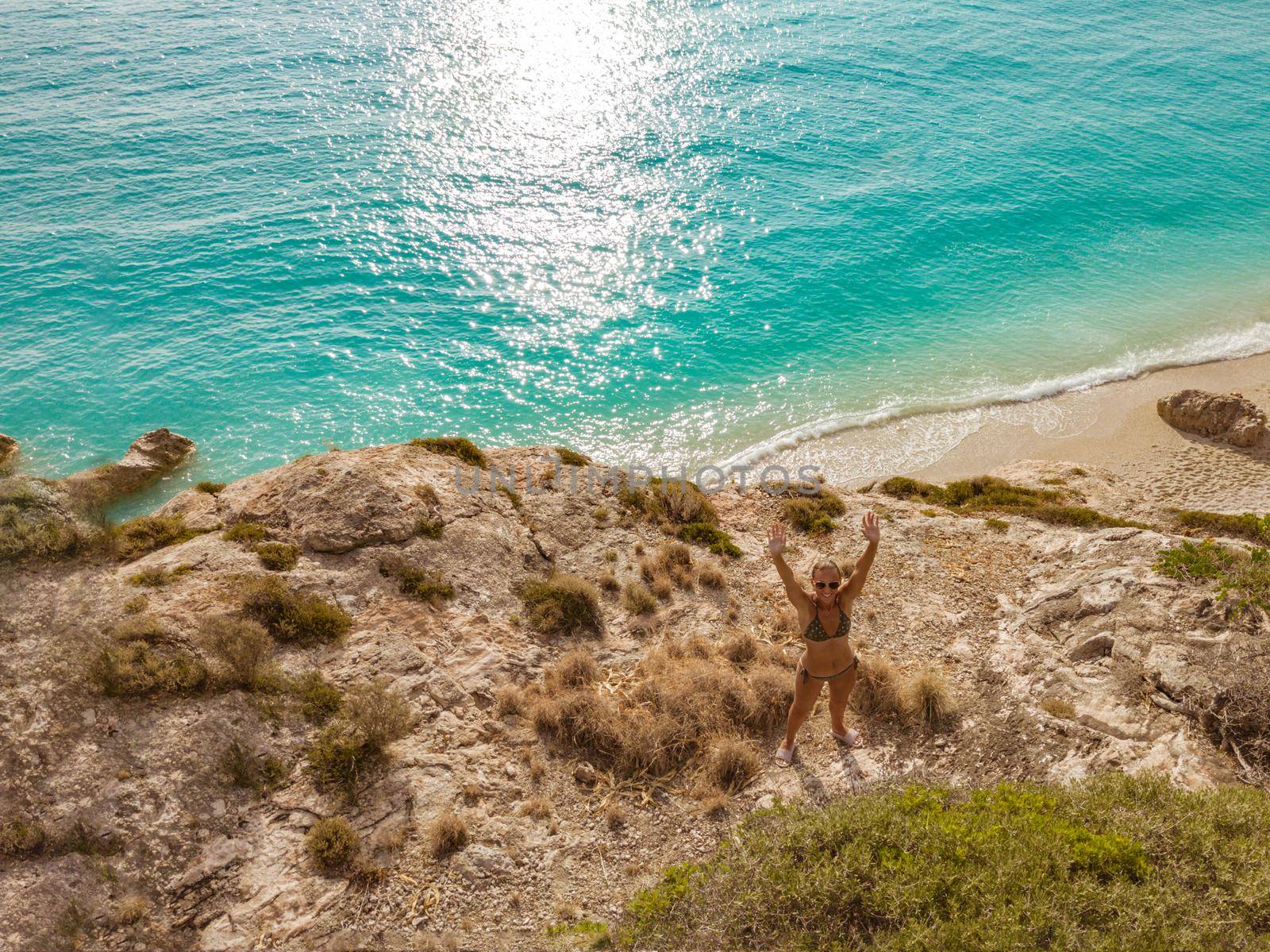 Top view of a beautiful woman enjoying in sunbath on the beach in a bikini, relaxing near clear sea water. She is waving and looking at camera.